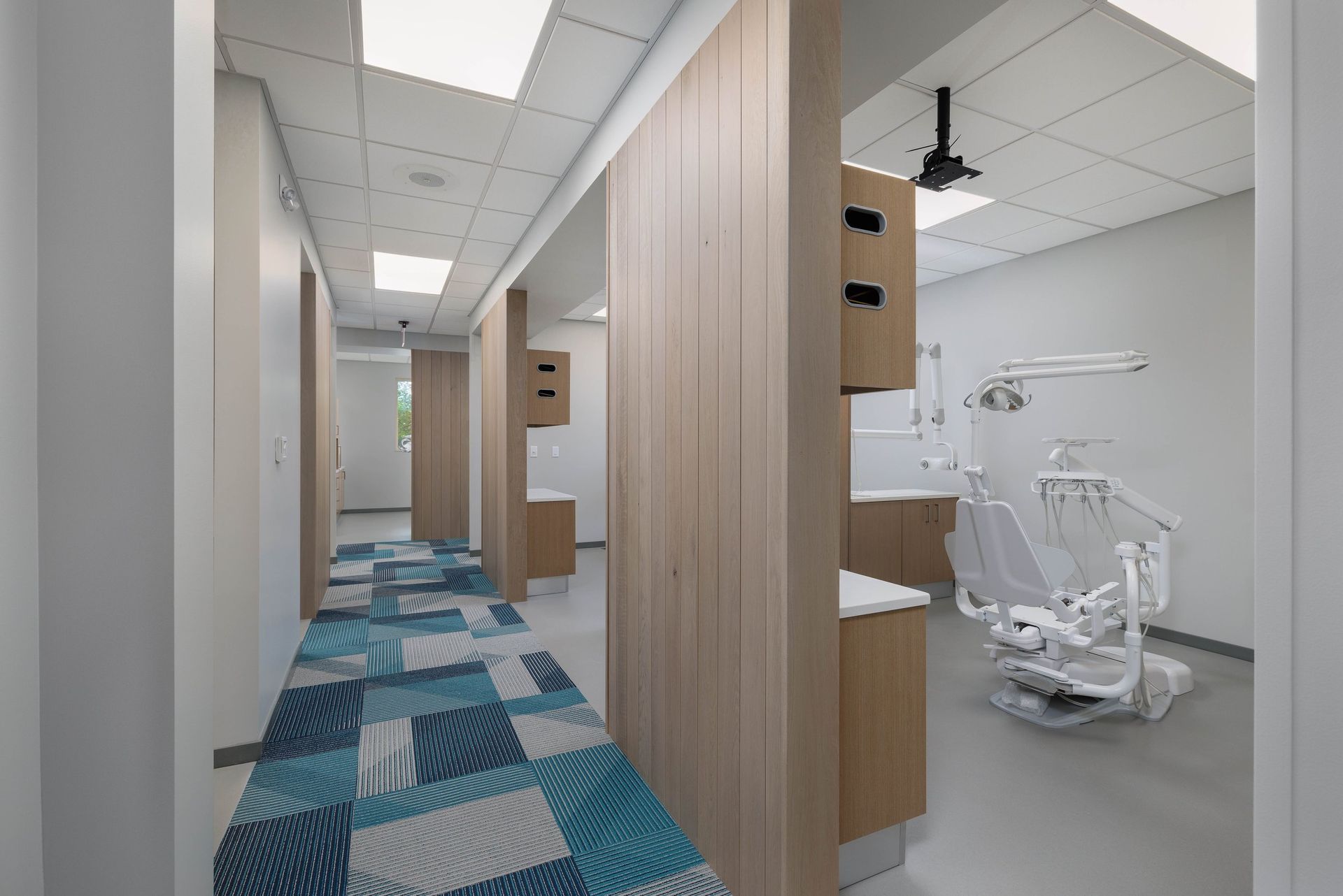 A brightly lit dental office hallway with blue patterned carpet leads past wooden partitions into a clinical treatment room.