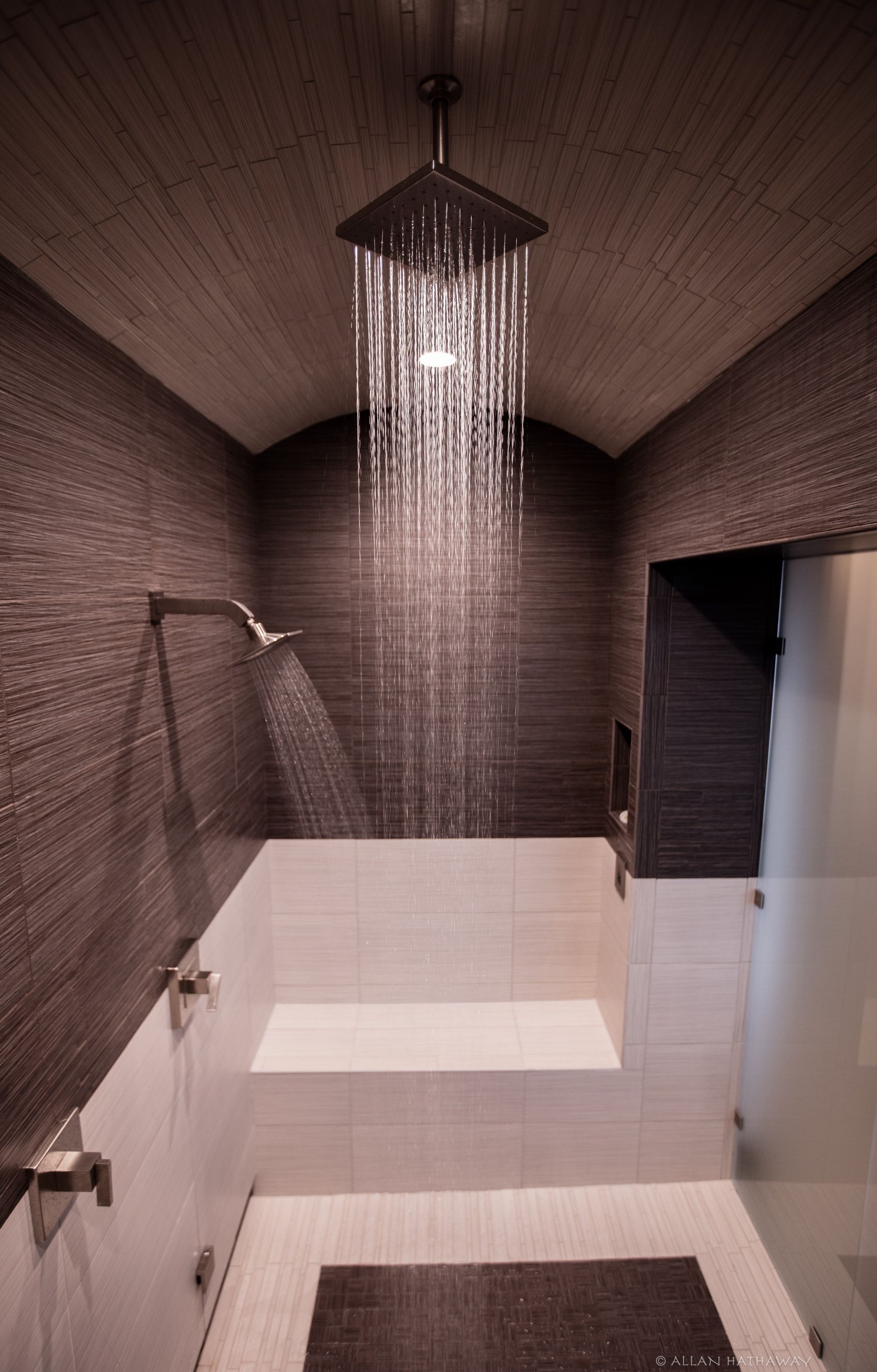 A modern shower with dark textured walls, white tile, a built-in bench, and both overhead and wall-mounted showerheads.