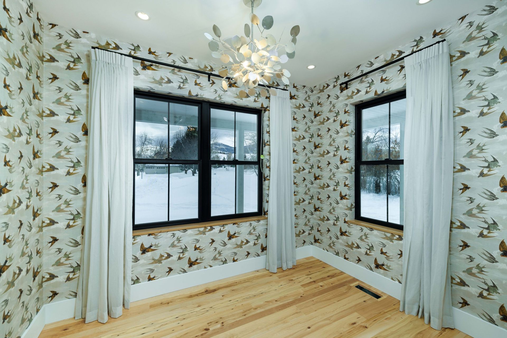 A room with bird-patterned wallpaper, two windows with light curtains, wooden floors, and a leaf-shaped chandelier.