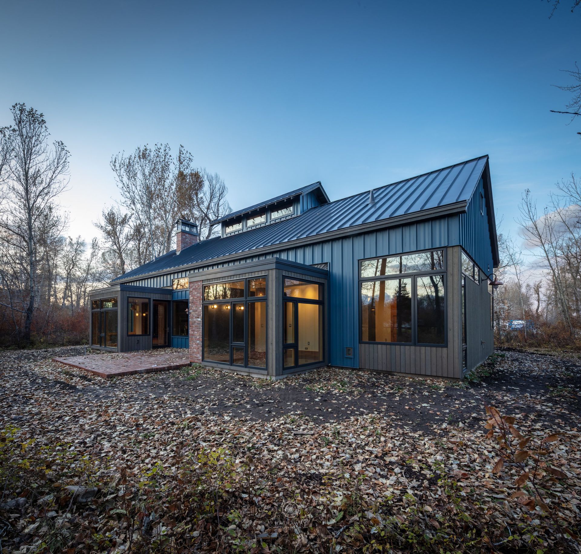 A modern house with blue metal siding and a glass sunroom, set in a wooded area covered in fallen autumn leaves.