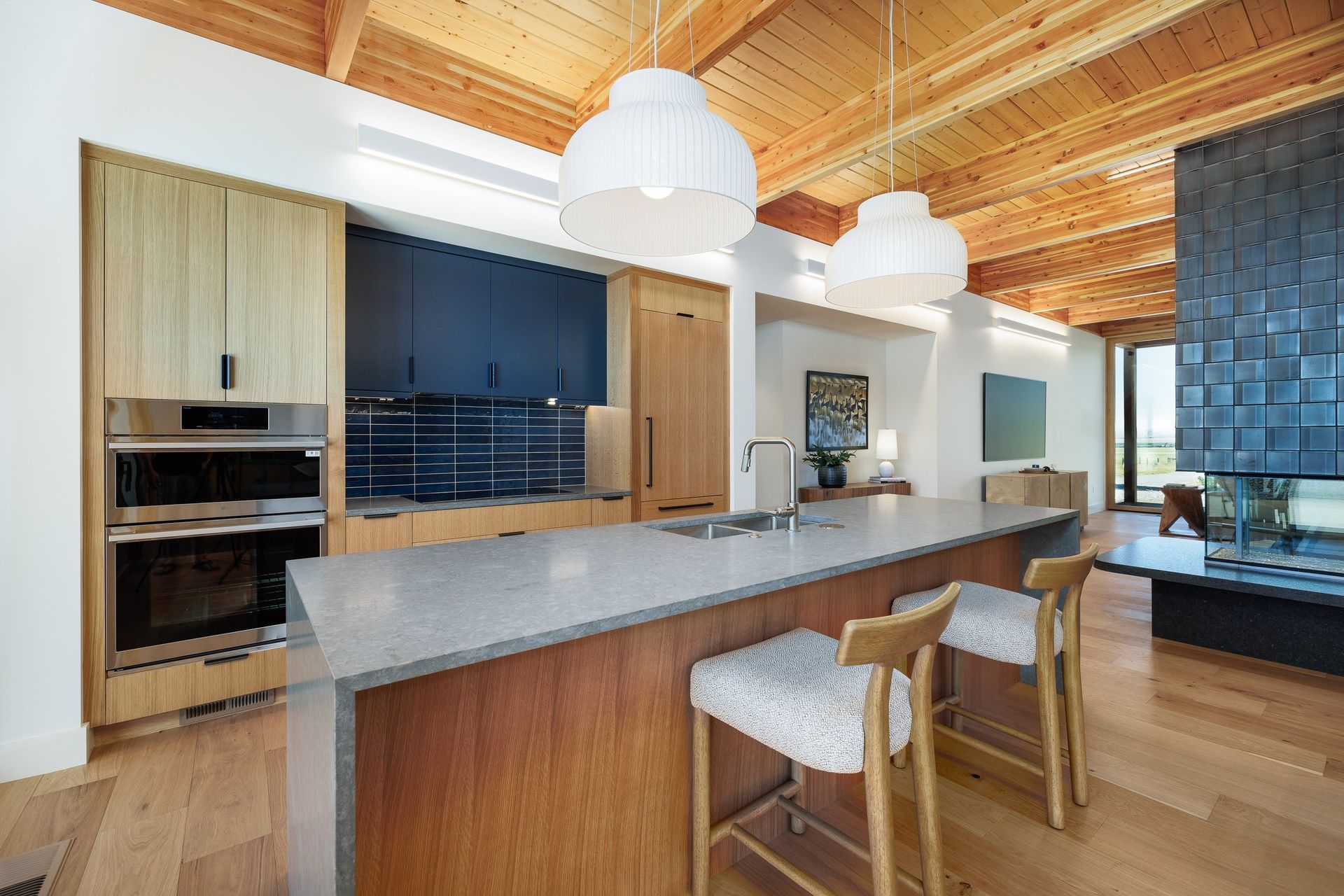Modern kitchen with a gray island, wooden cabinets, dark blue backsplash, dual wall ovens, and two pendant lights.