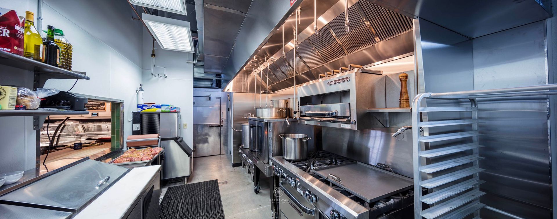 A clean, professional stainless steel commercial kitchen featuring a stove, range hood, prep tables, and shelving.