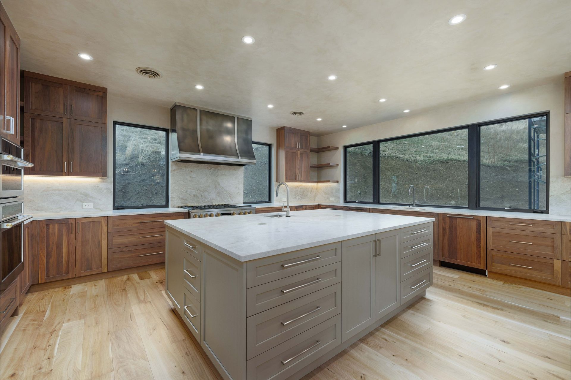 A modern kitchen with a large gray island, walnut cabinetry, white countertops, and a wide window with a black frame.