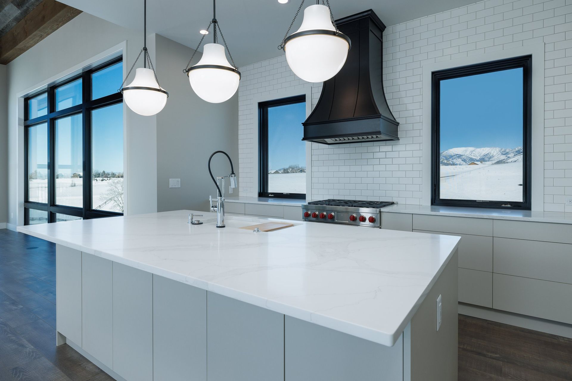 A modern kitchen with a large white island, gray cabinets, black accents, and large windows overlooking a snowy landscape.