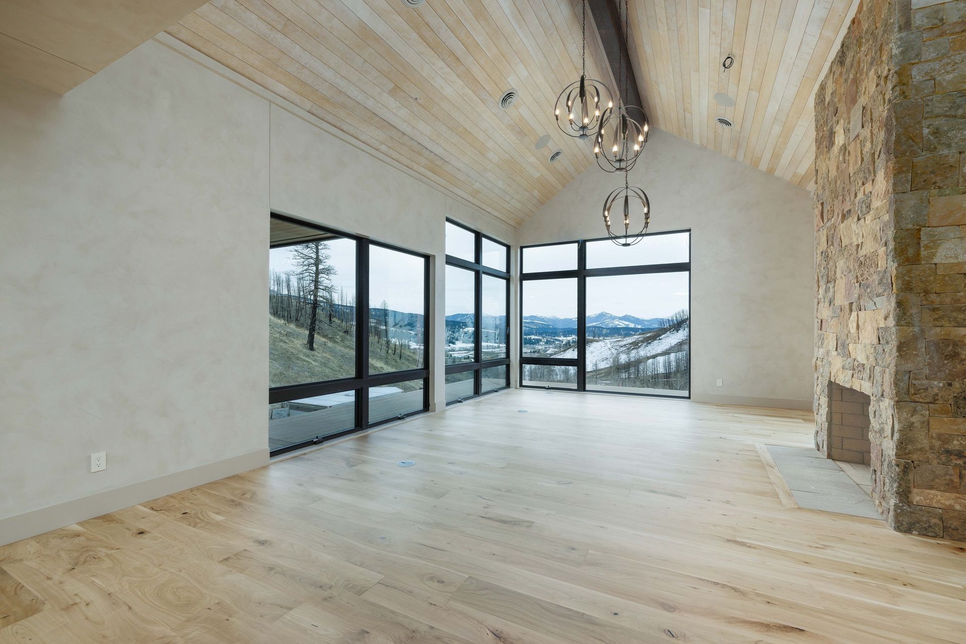 A spacious, modern room with light wood floors, a vaulted wood ceiling, floor-to-ceiling windows, and a stone fireplace.