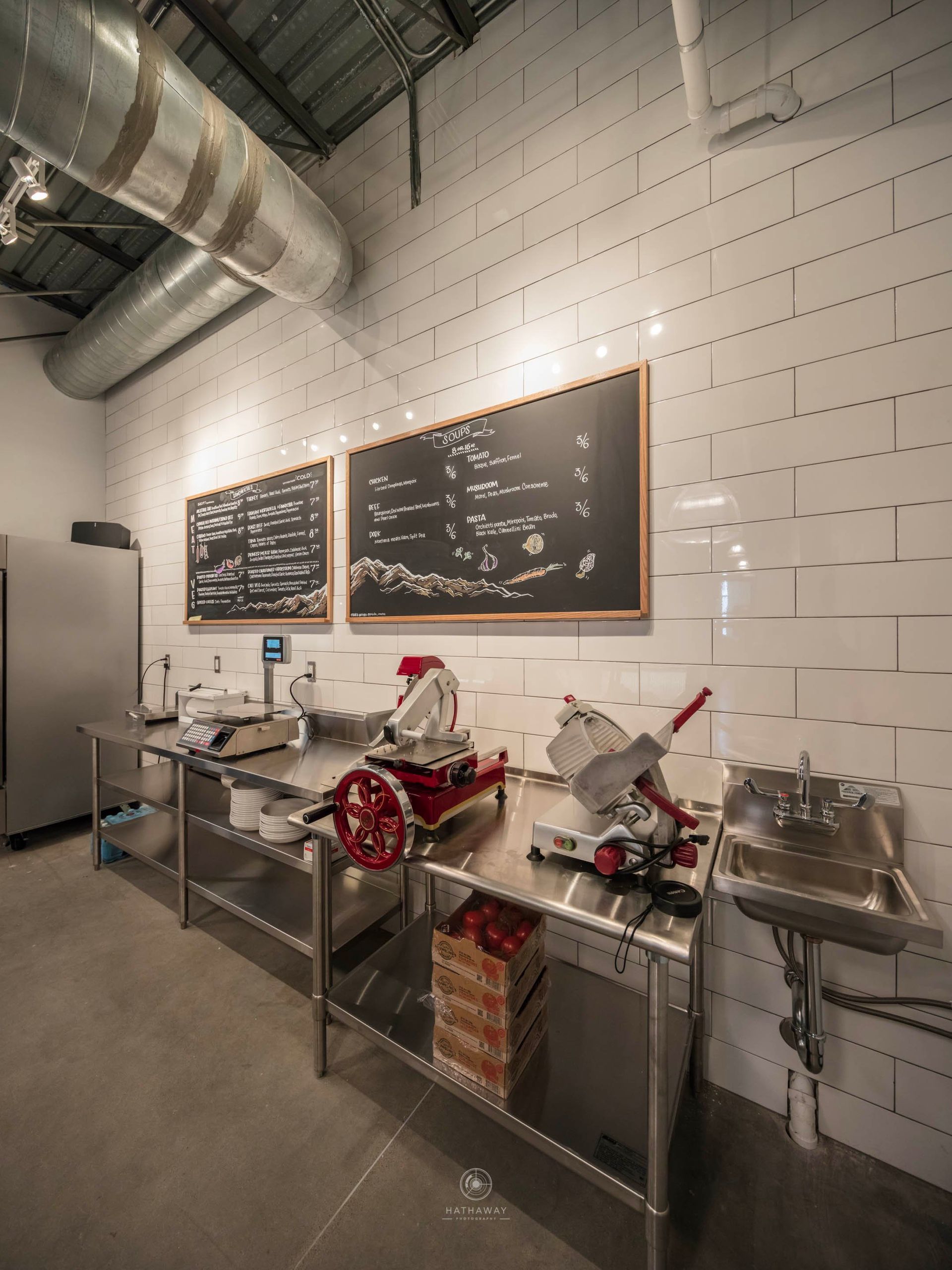 Professional kitchen workspace with industrial food slicers, stainless steel counters, and chalkboards on a tiled wall.