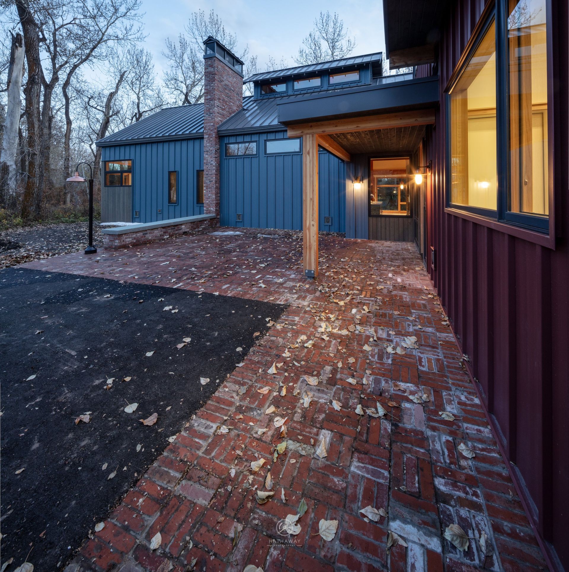 A blue and burgundy home with a brick patio and a black driveway in a wooded area during twilight.