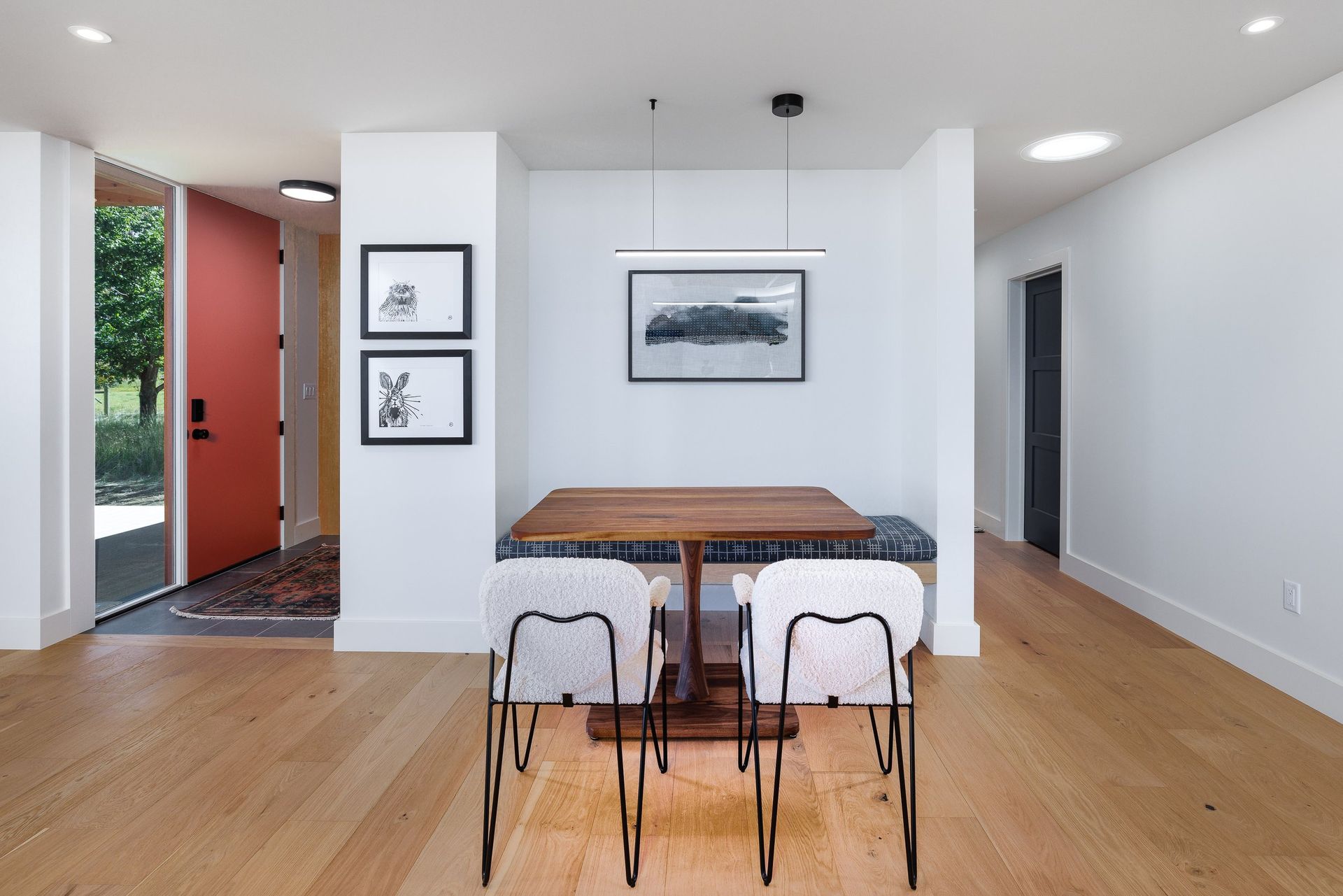 A dining nook with a wooden table, two chairs, and a bench, featuring white walls, hardwood floors, and a red door.
