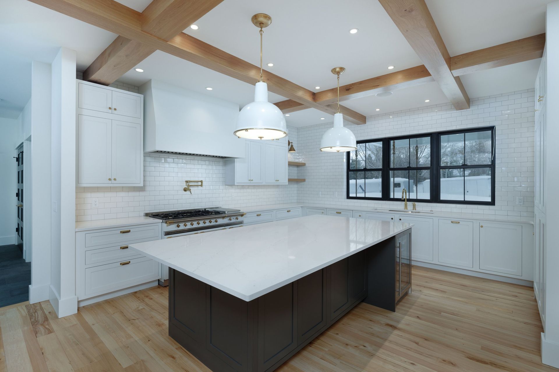 A modern kitchen featuring white cabinets, a large dark island, light wood floors, exposed ceiling beams, and a brick wall.