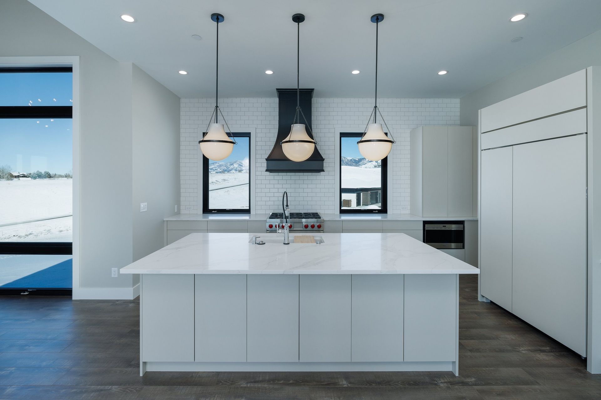 A modern, minimalist kitchen featuring a large white island, grey cabinetry, a black range hood, and three pendant lights.