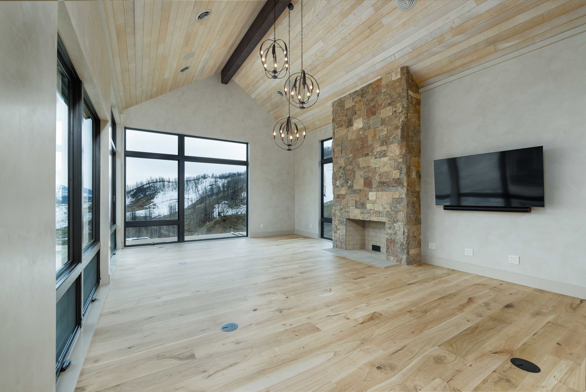 A modern living room with light wood floors, a tall stone fireplace, a flat-screen TV, and large windows with mountain views.