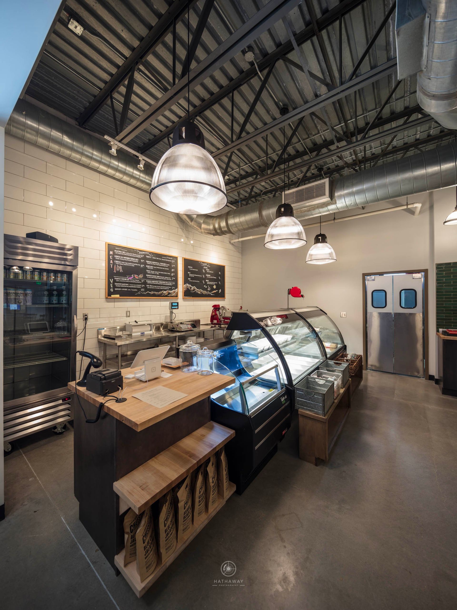 A modern café interior with industrial ceiling, hanging pendant lights, service counter, and a refrigerated display case.
