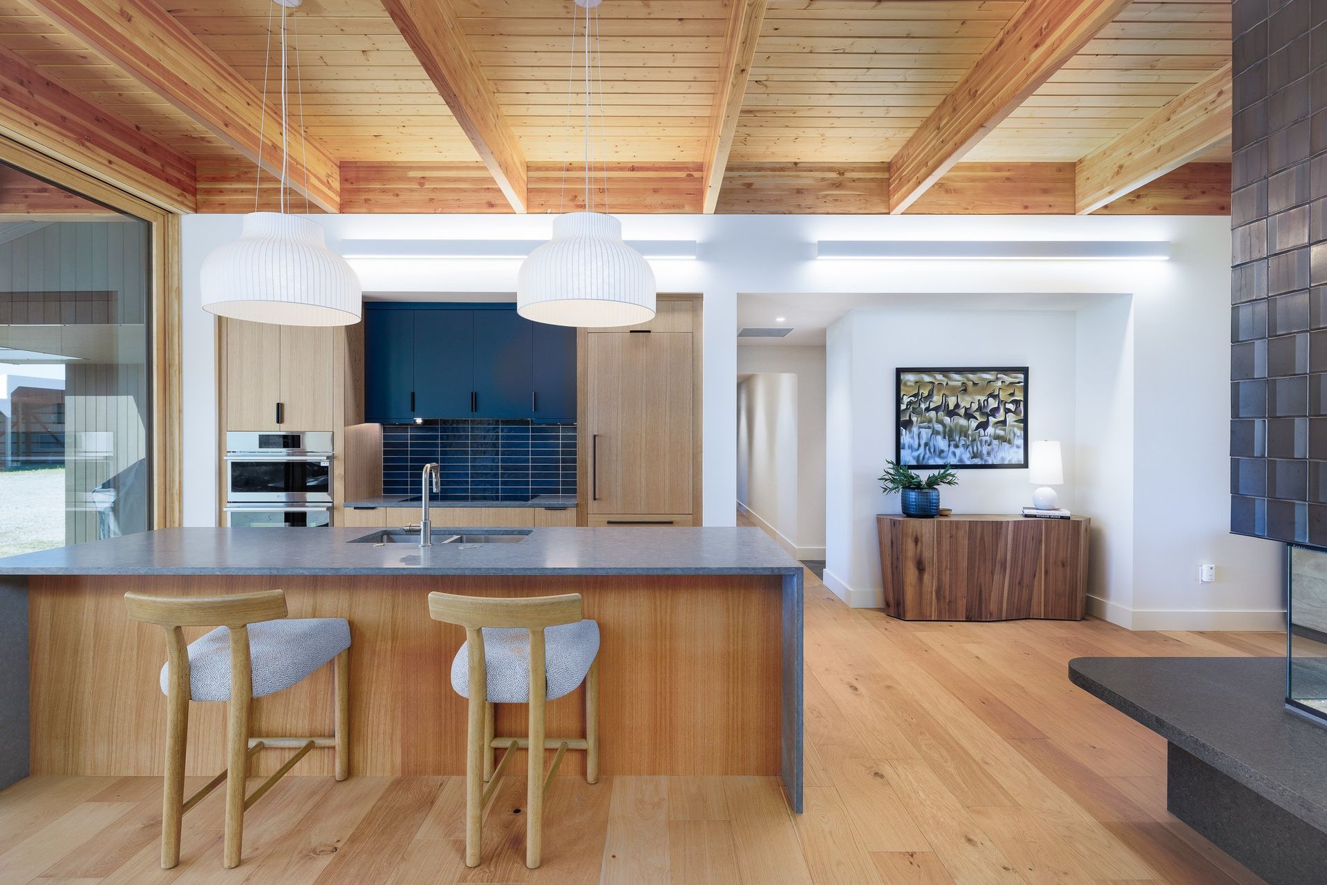 Modern kitchen with a gray island, wooden cabinets, bar stools, and vaulted timber ceilings.