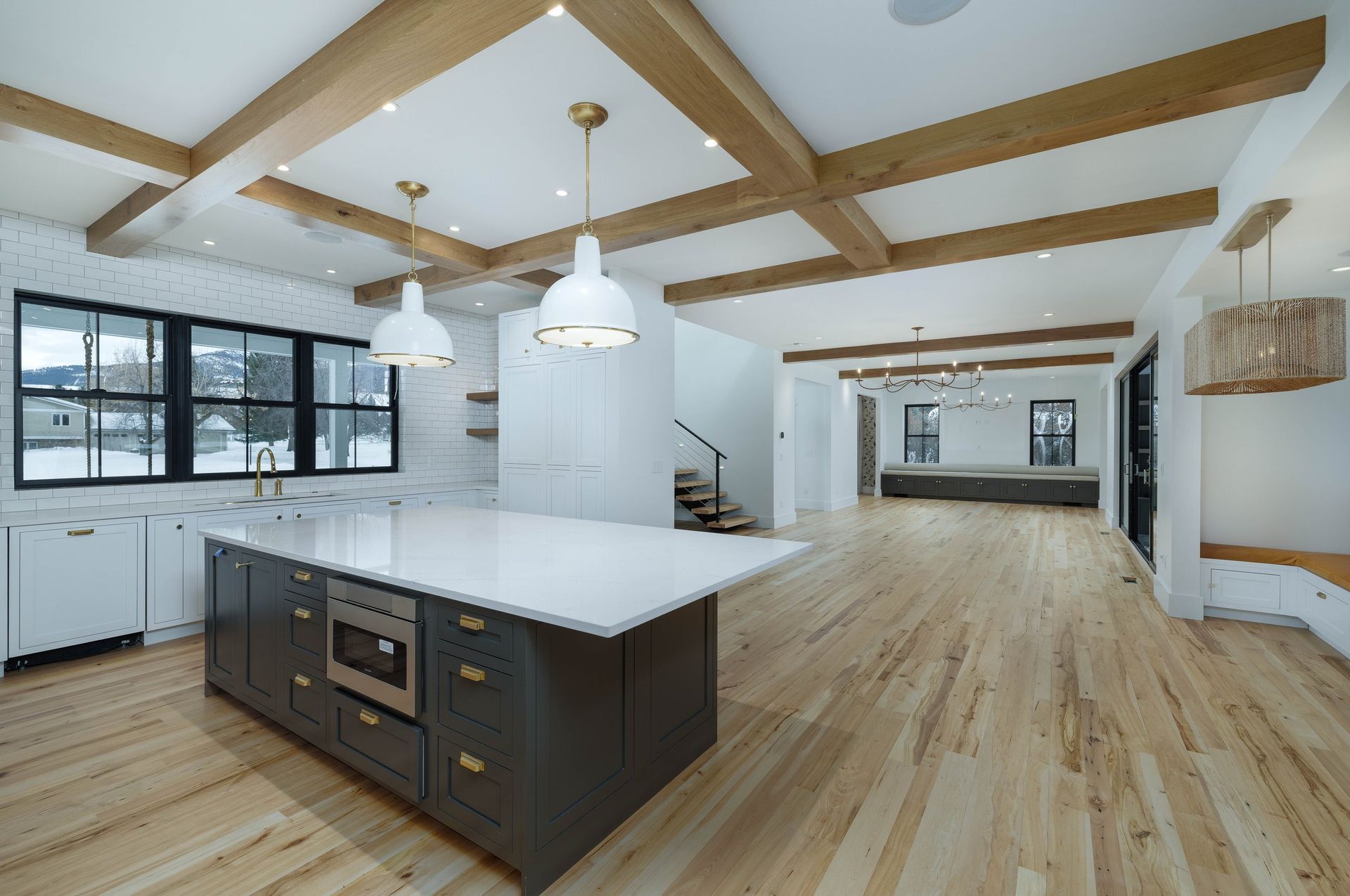 A kitchen with a dark island, white counters, and wood-beamed ceilings, opening into a spacious room with wood floors.