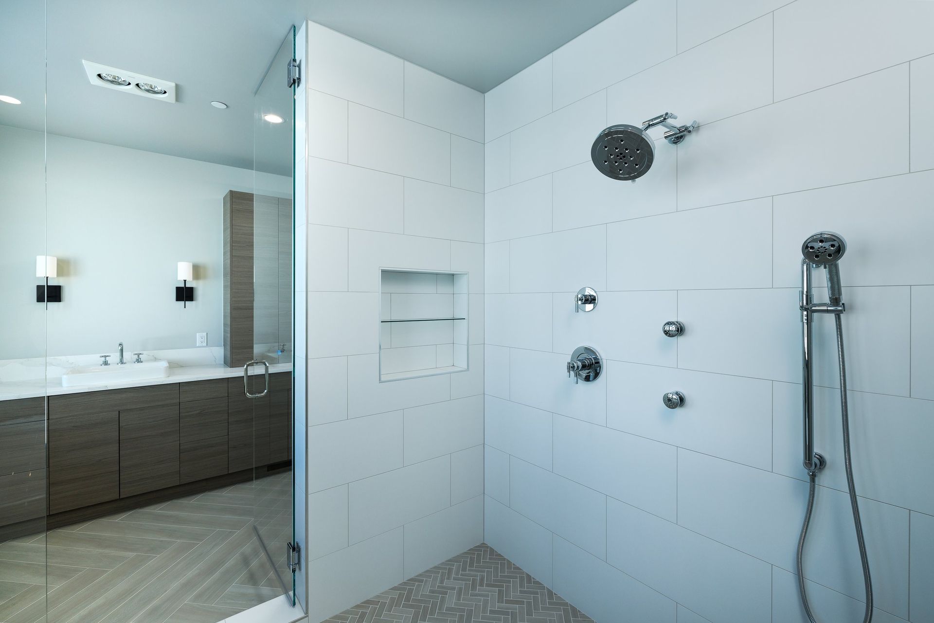 A modern bathroom featuring a glass-enclosed shower with white tiled walls, multiple shower heads, and a wood vanity.