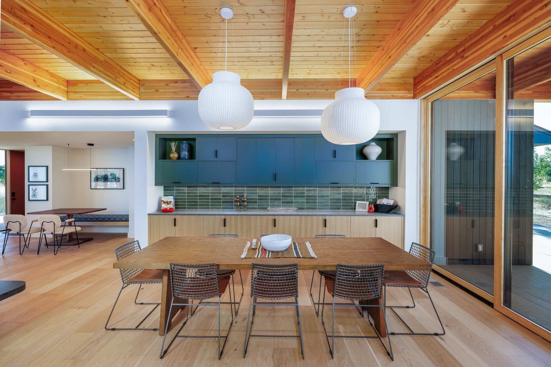 A modern dining room featuring a wooden table, wire chairs, blue cabinets, and globe pendant lights under a wood ceiling.