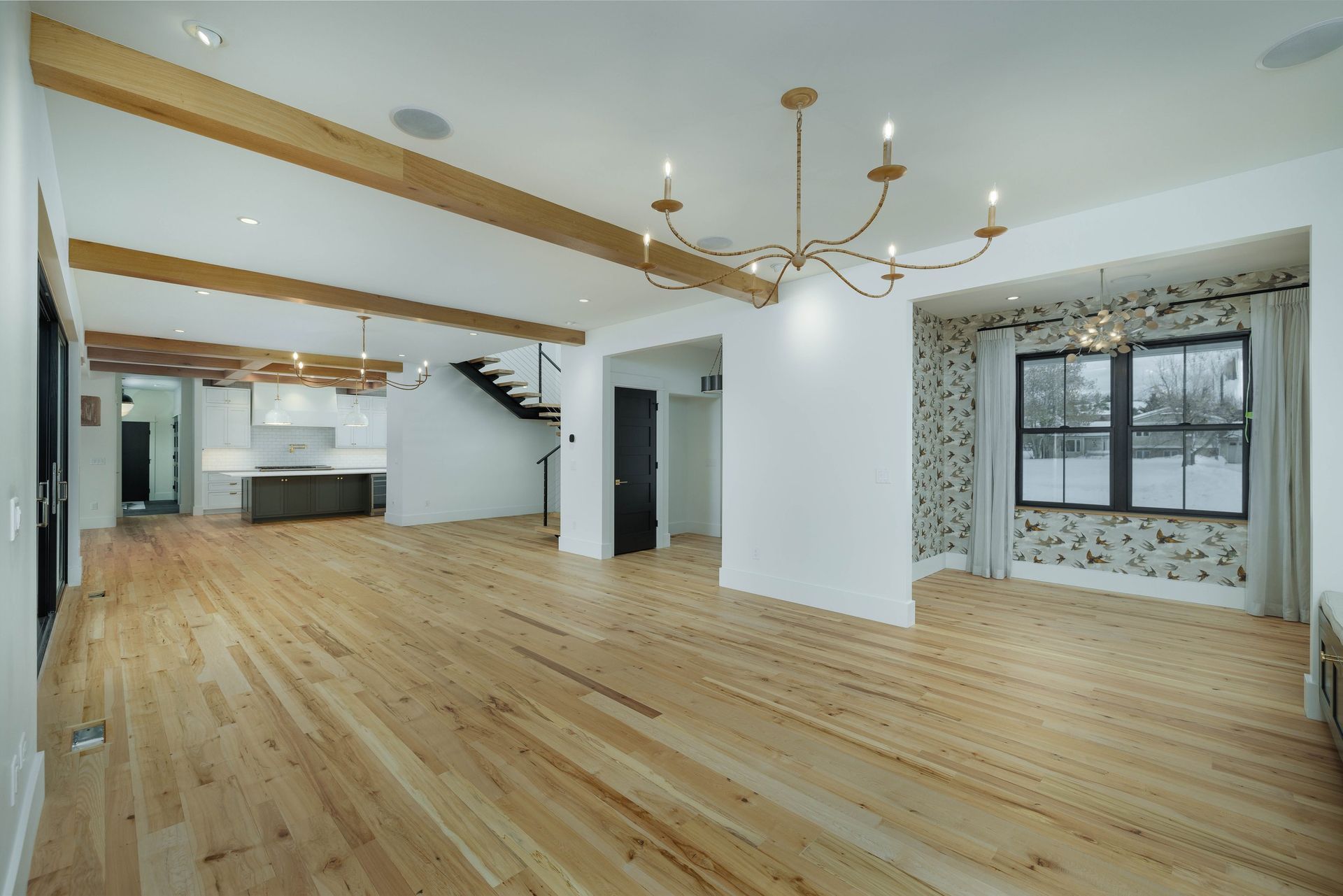 A spacious, modern open-plan room with light wood floors, white walls, exposed wooden ceiling beams, and a chandelier.