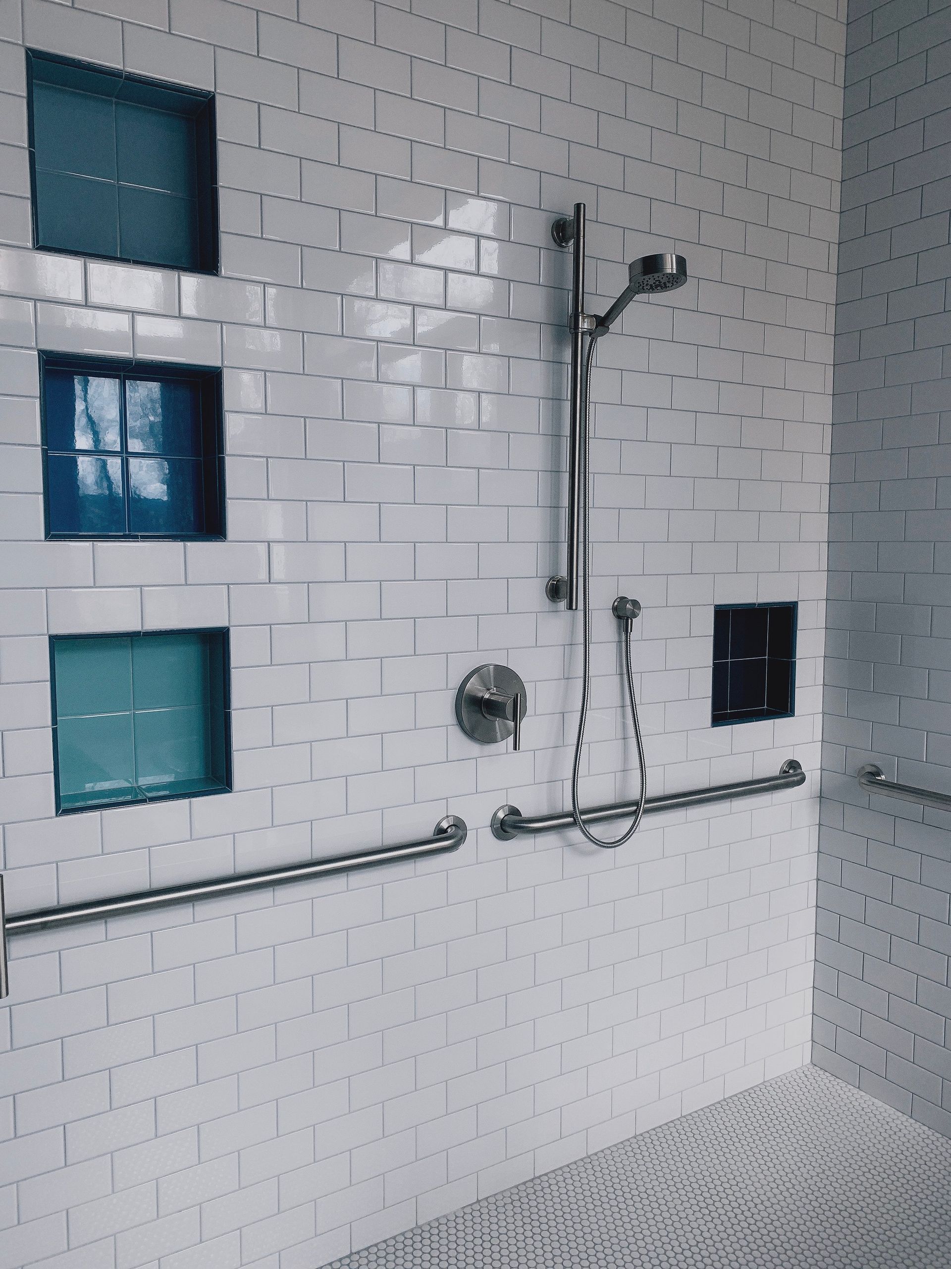 A tiled, accessible shower with a handheld sprayer, wall-mounted grab bars, and blue recessed accent niches.