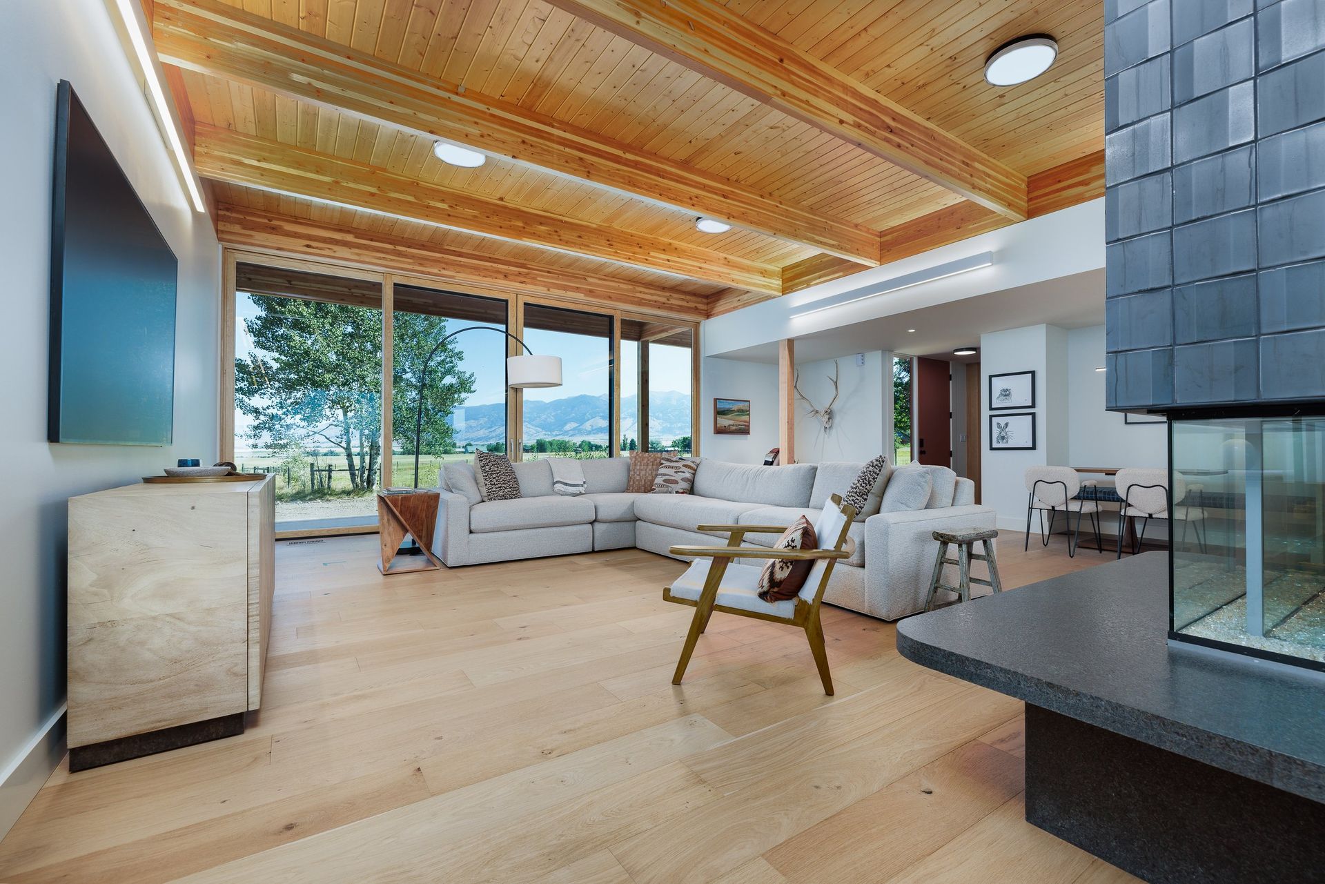 Modern living room with a sectional sofa, wooden ceiling, and a large window looking out to a landscape.