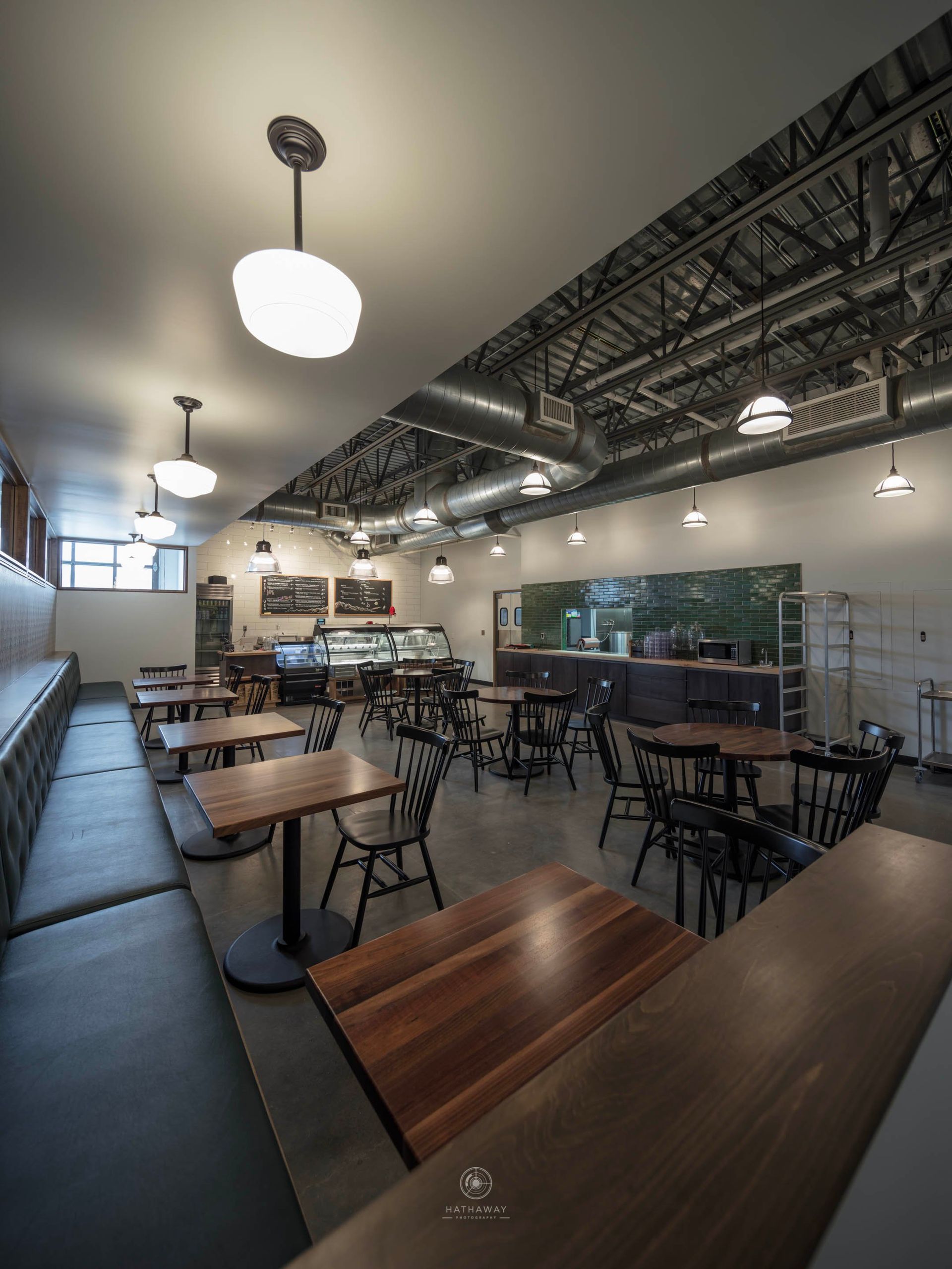 A wide-angle view of a restaurant interior with wooden tables, black chairs, a long bench, and exposed ceiling ductwork.