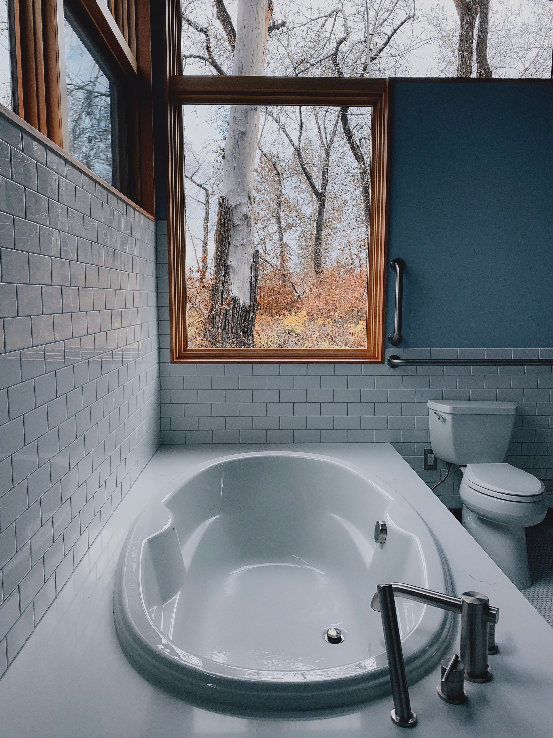 A modern bathroom featuring a white bathtub, tiled walls, and a large window looking out onto a forest in autumn.