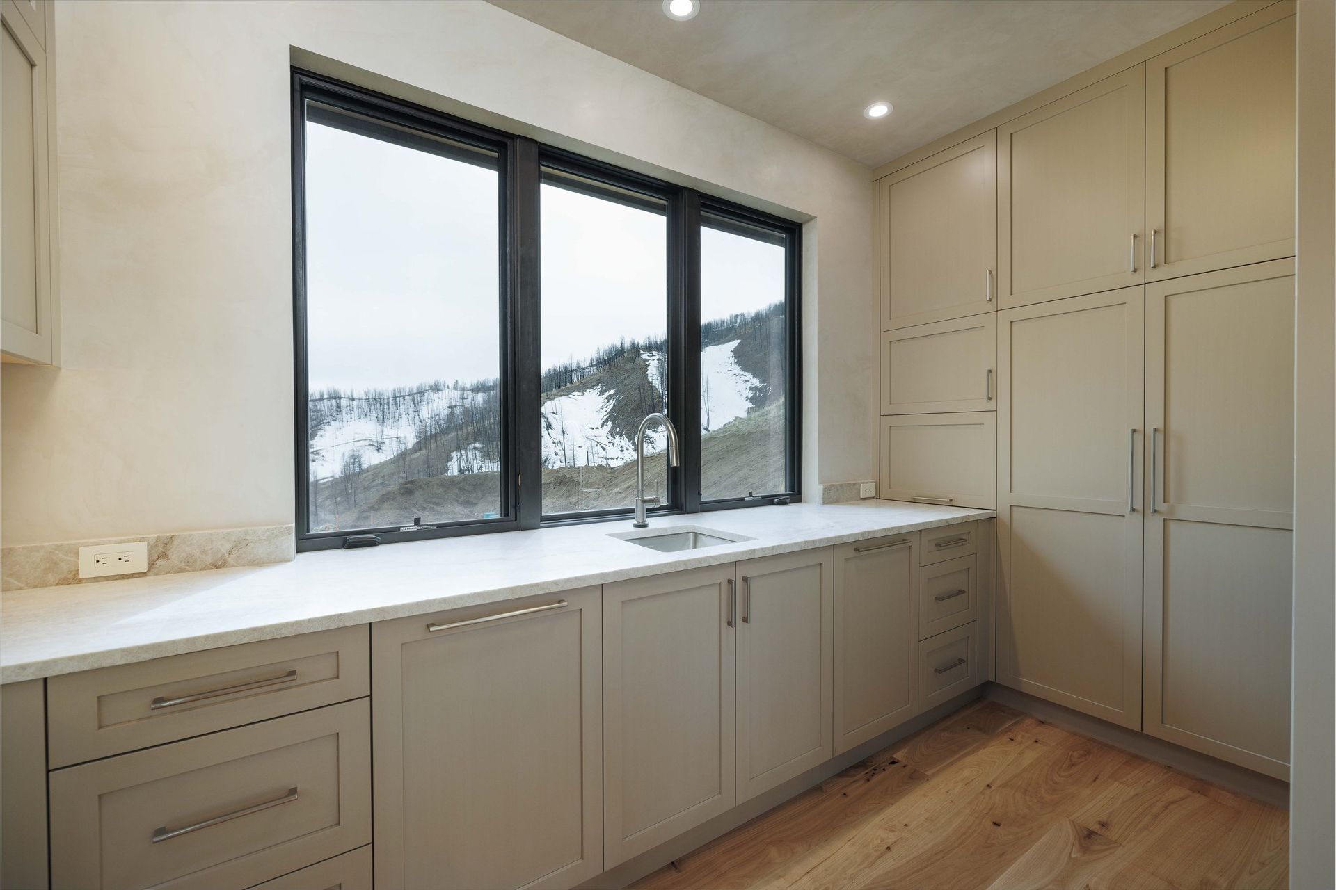 A modern kitchen featuring light beige cabinetry, white countertops, and a large window overlooking a snowy mountainside.