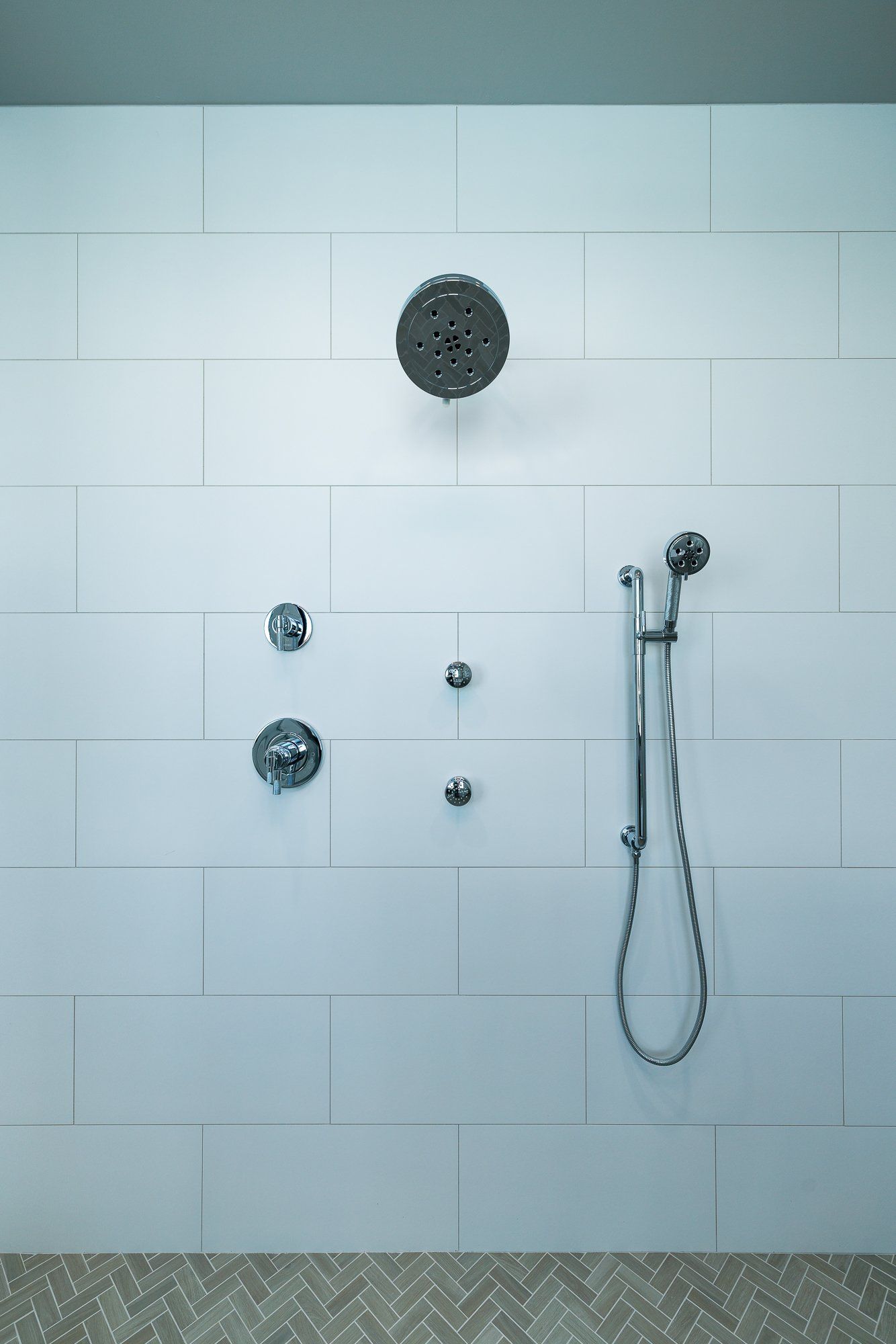 A white tiled shower wall with a round overhead shower head, a handheld sprayer on a rail, and two chrome control knobs.