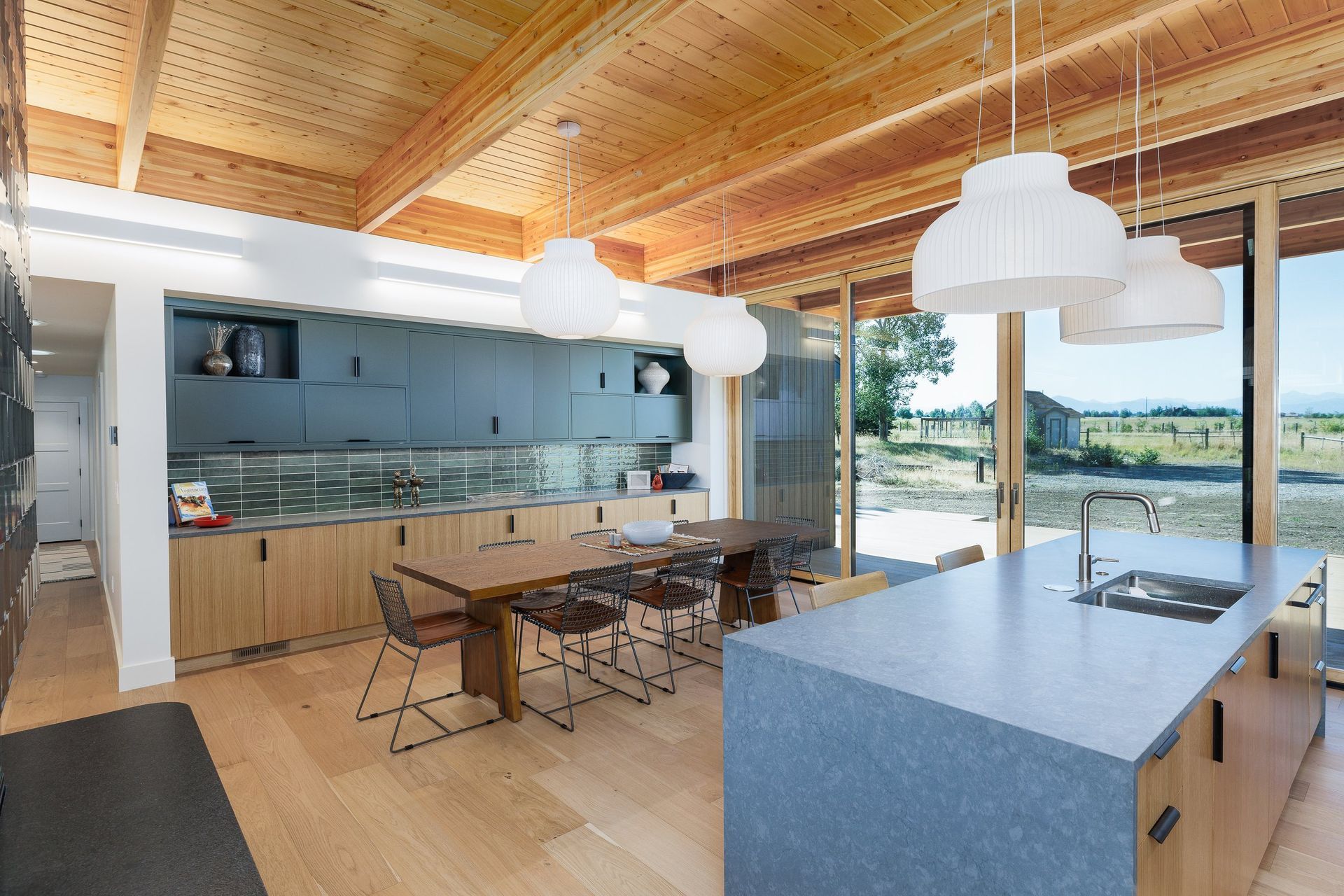 A modern, open-plan kitchen with wood ceilings, navy blue cabinetry, a long dining table, and a gray-topped kitchen island.