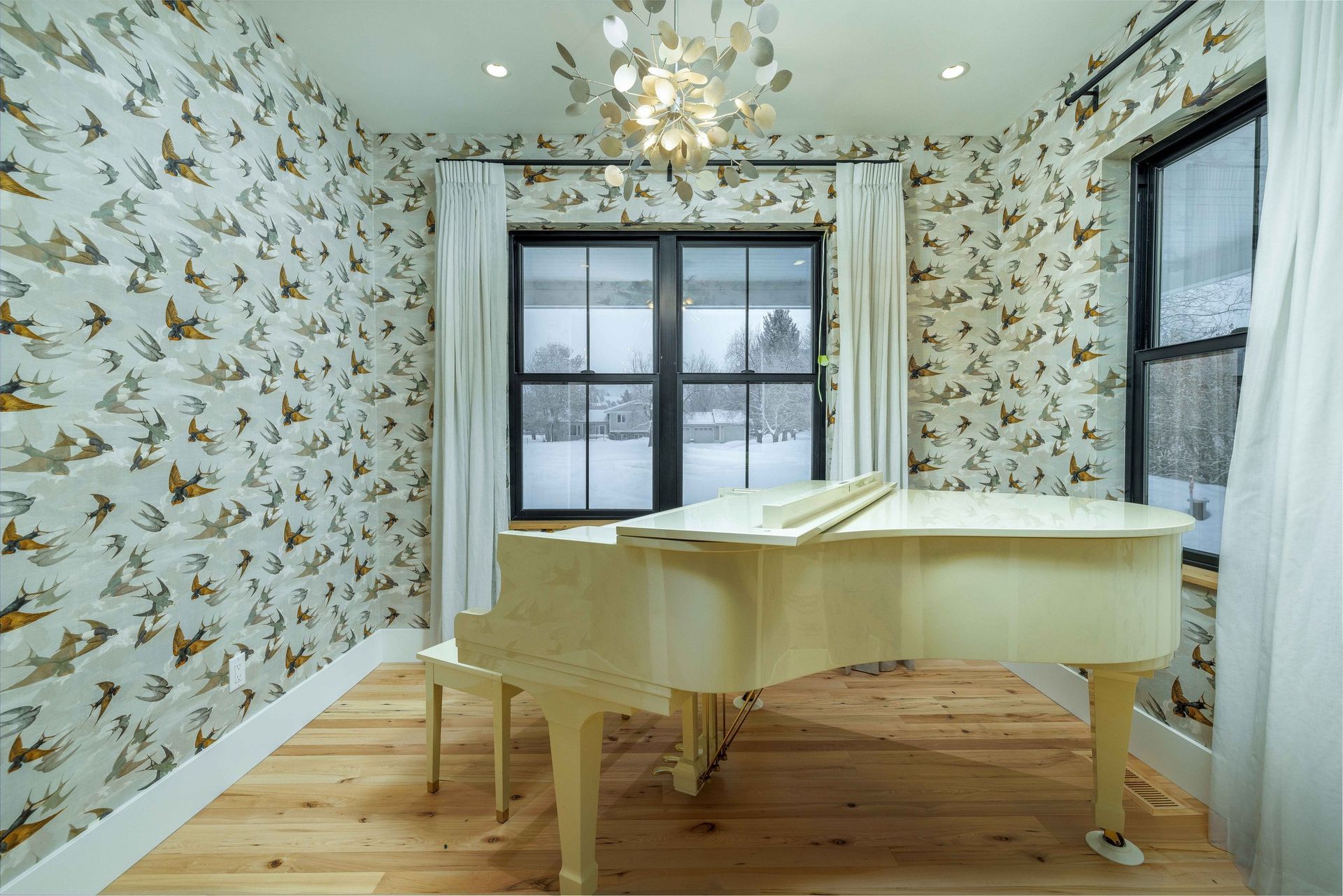 A white grand piano sits on a hardwood floor in a room with bird-patterned wallpaper and a window overlooking snow.