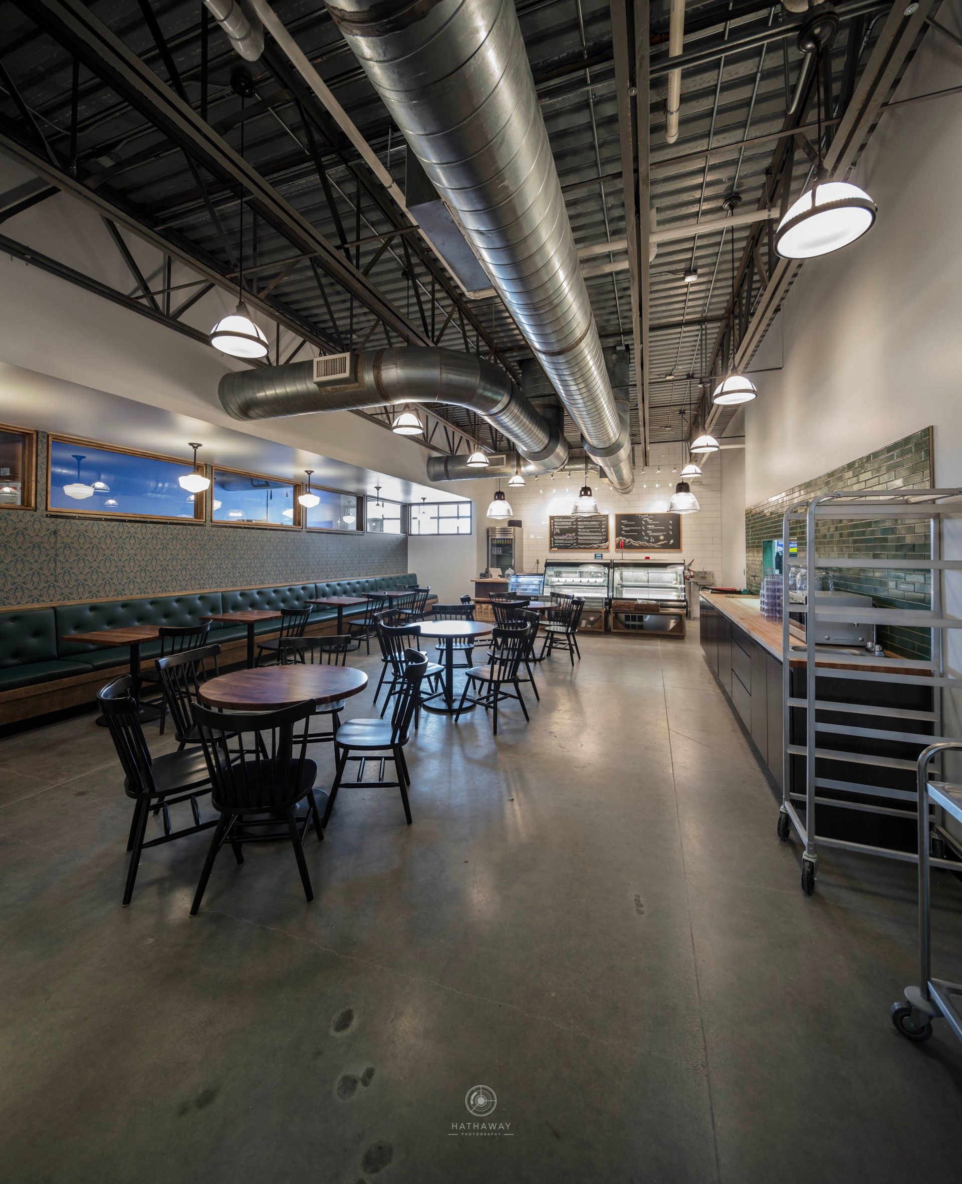 Empty restaurant interior with round tables, chairs, a long bench, industrial ceiling ducts, and a metal service rack.