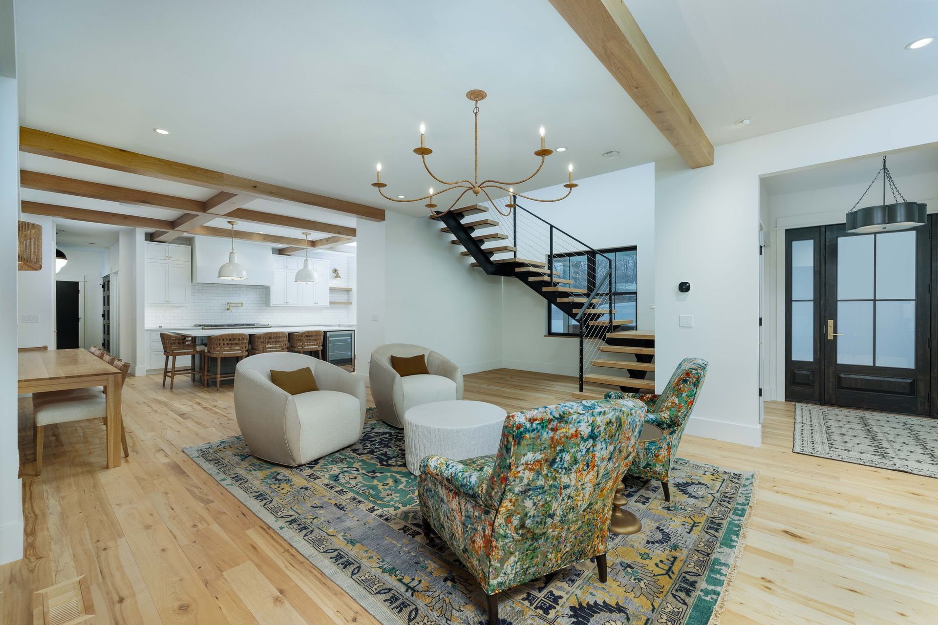 A modern, open-concept living area with white walls, wood ceiling beams, a chandelier, and a dark staircase.