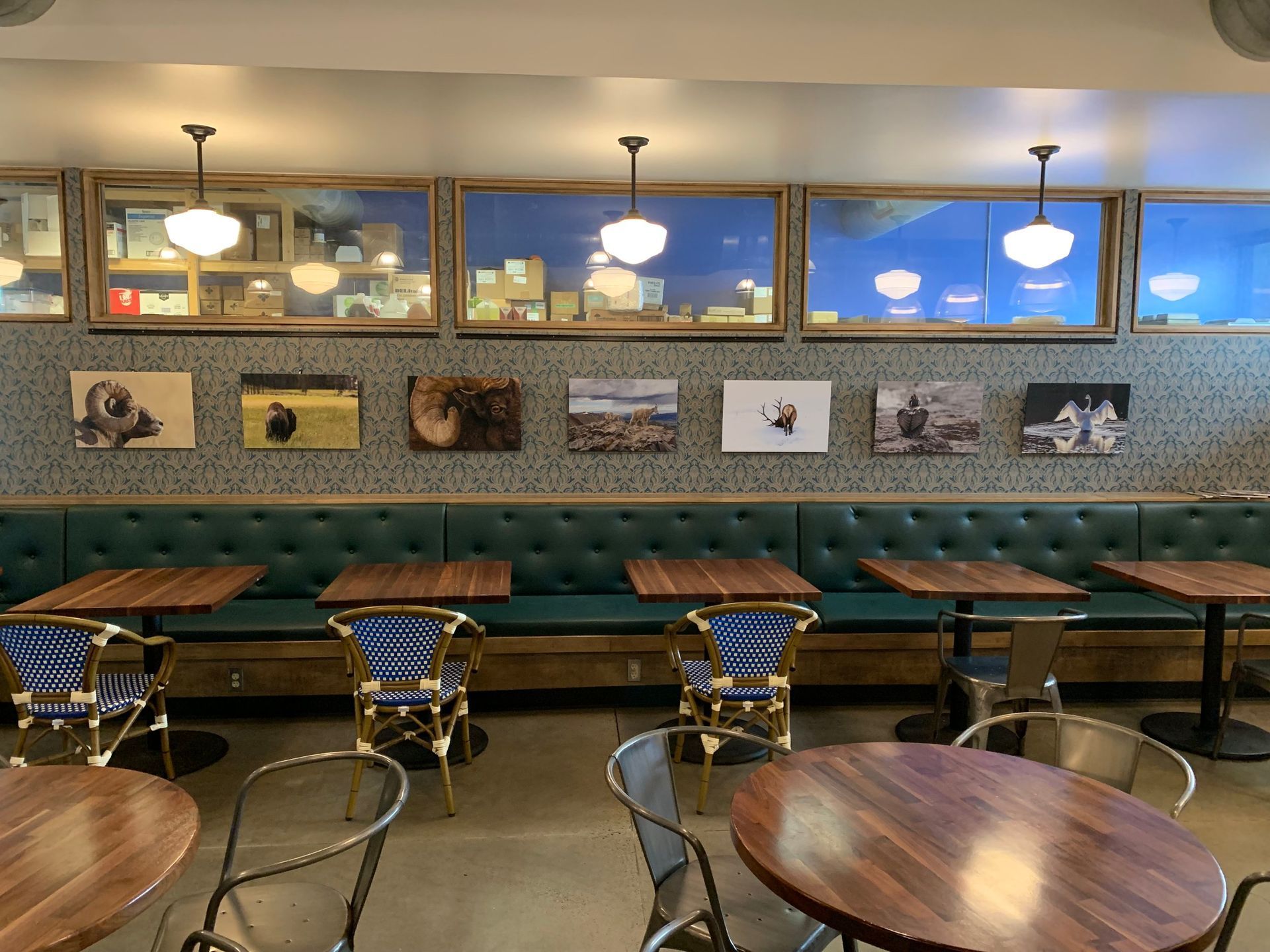 A restaurant interior with a tufted teal bench, wooden tables, bistro chairs, and framed artwork on a patterned wall.