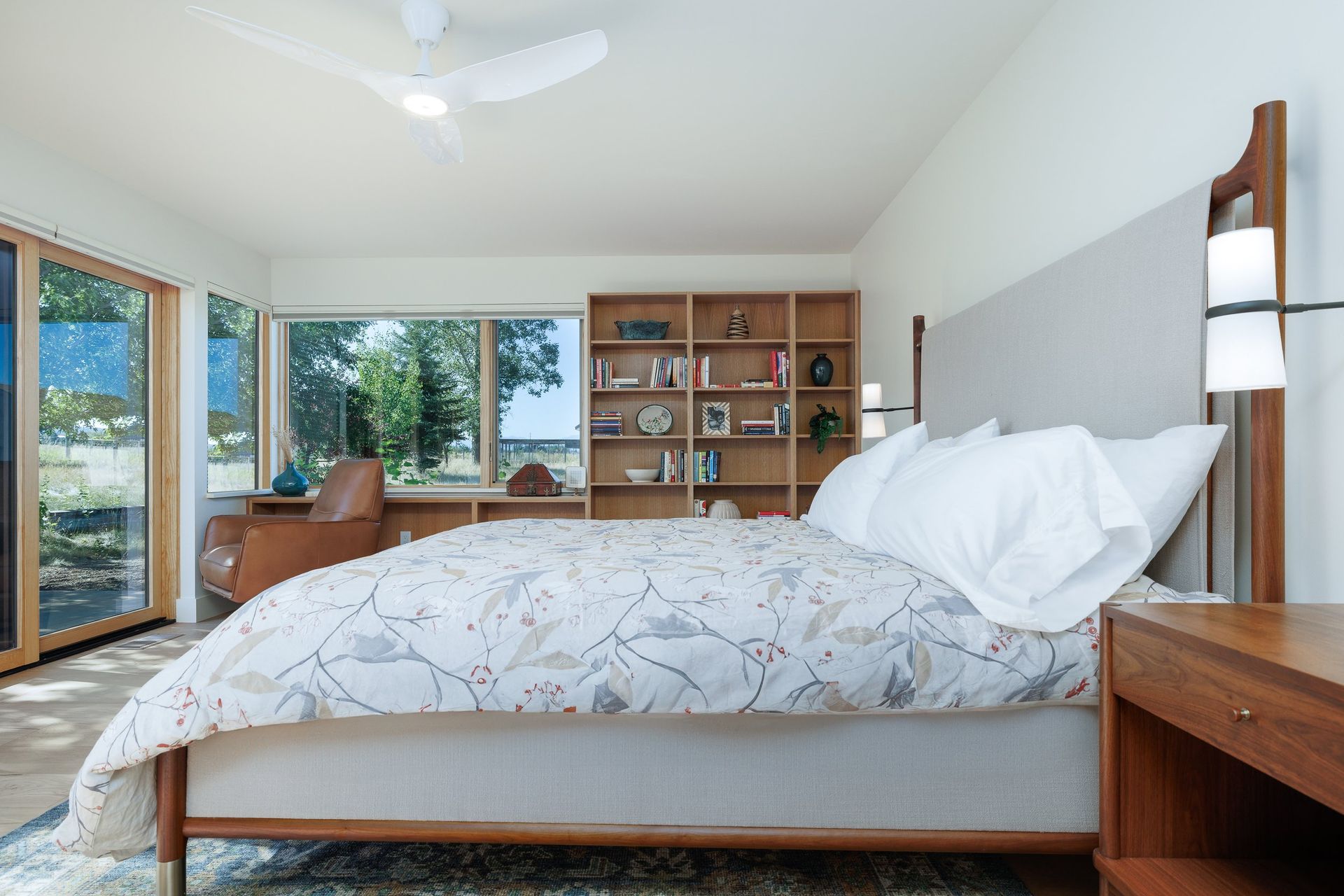 A modern bedroom features a bed with a floral duvet, a wooden headboard, a large bookshelf, and a chair by a glass door.