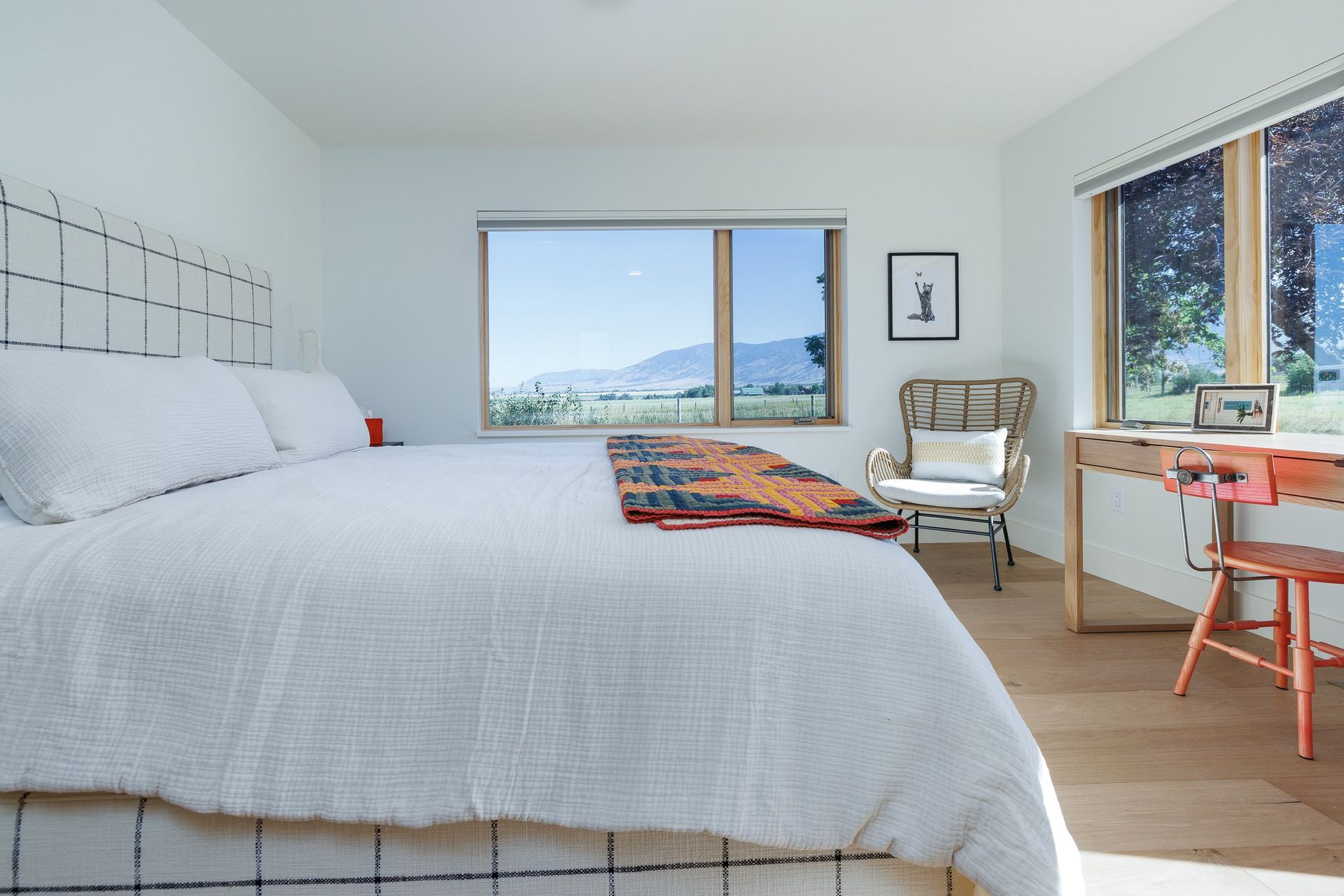 A bright, minimalist bedroom with a large bed, a scenic window, a patterned armchair, and a small wooden desk with a chair.