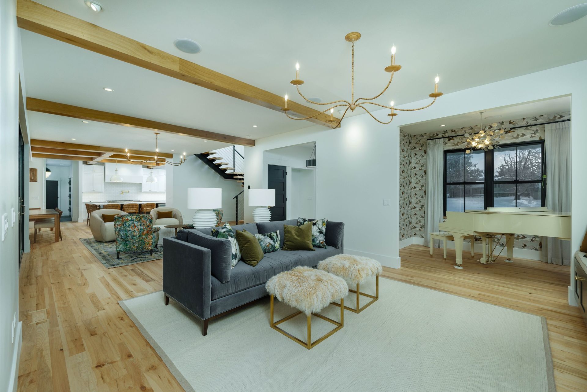 A bright, open-plan living room with light hardwood floors, a gray sofa, two white ottomans, and a grand piano.