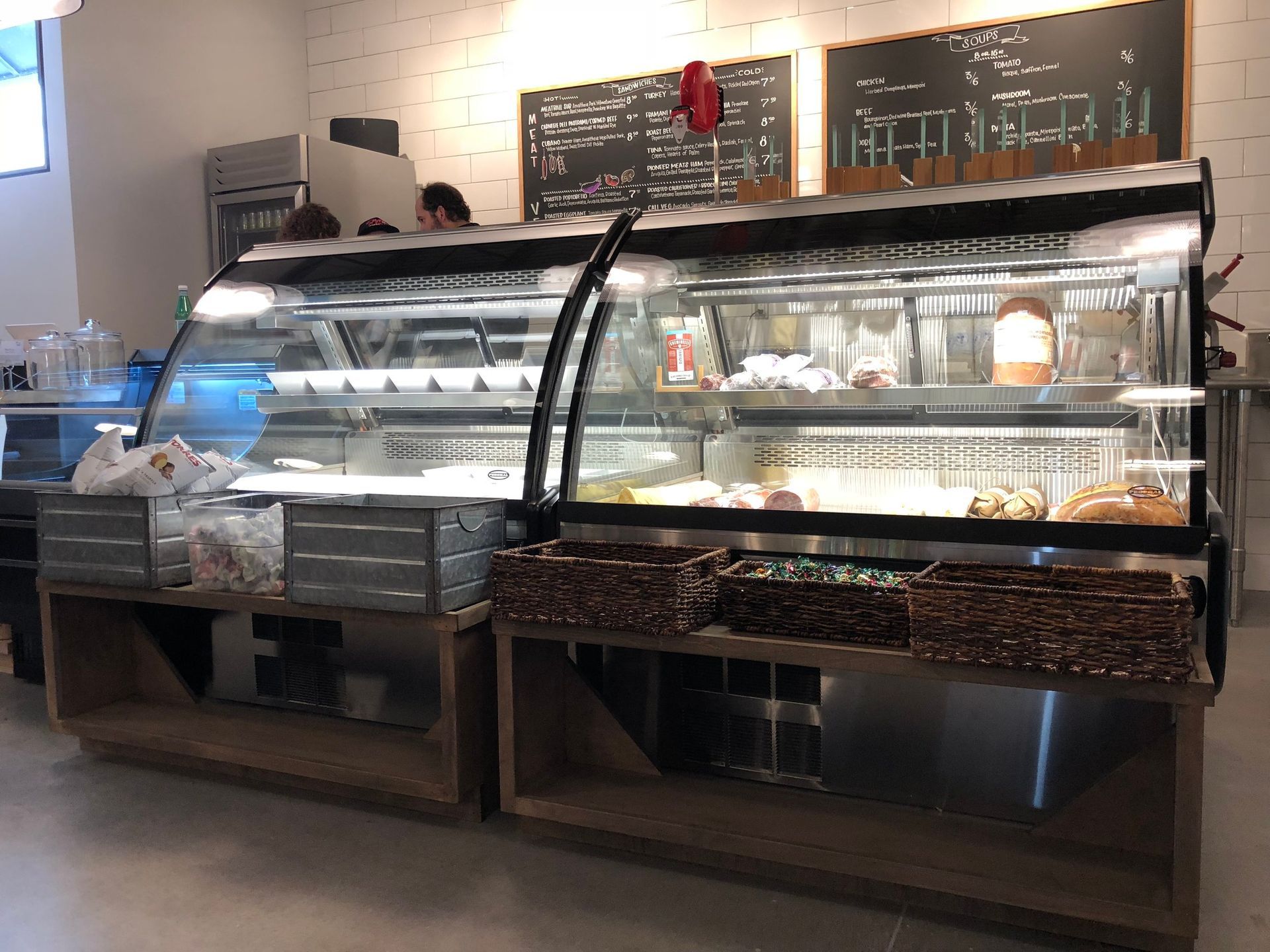Two refrigerated food display cases with dark wicker baskets on wooden counters in a cafe with menu chalkboards behind.