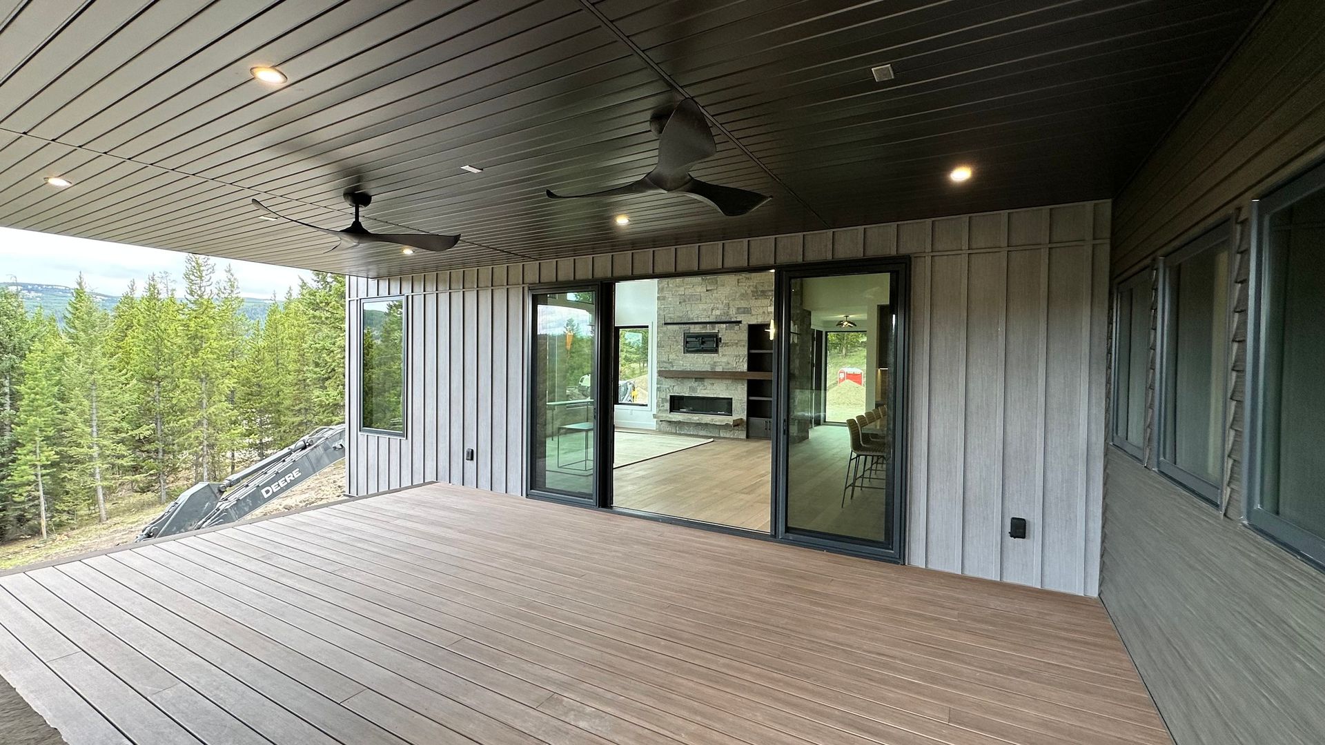 A covered outdoor deck with light brown wood decking, a dark paneled ceiling, and glass doors leading to a living area.