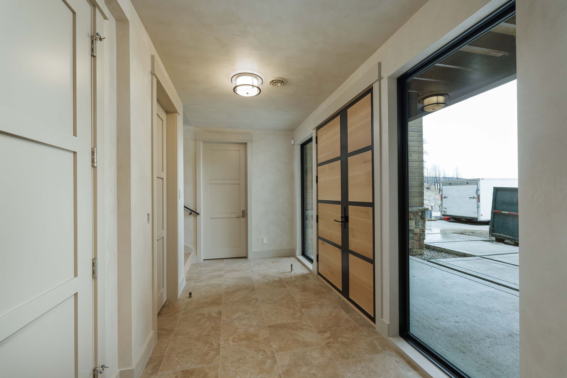 An indoor hallway with beige stone flooring, off-white walls, and doors leading to a bright, paved outdoor area.