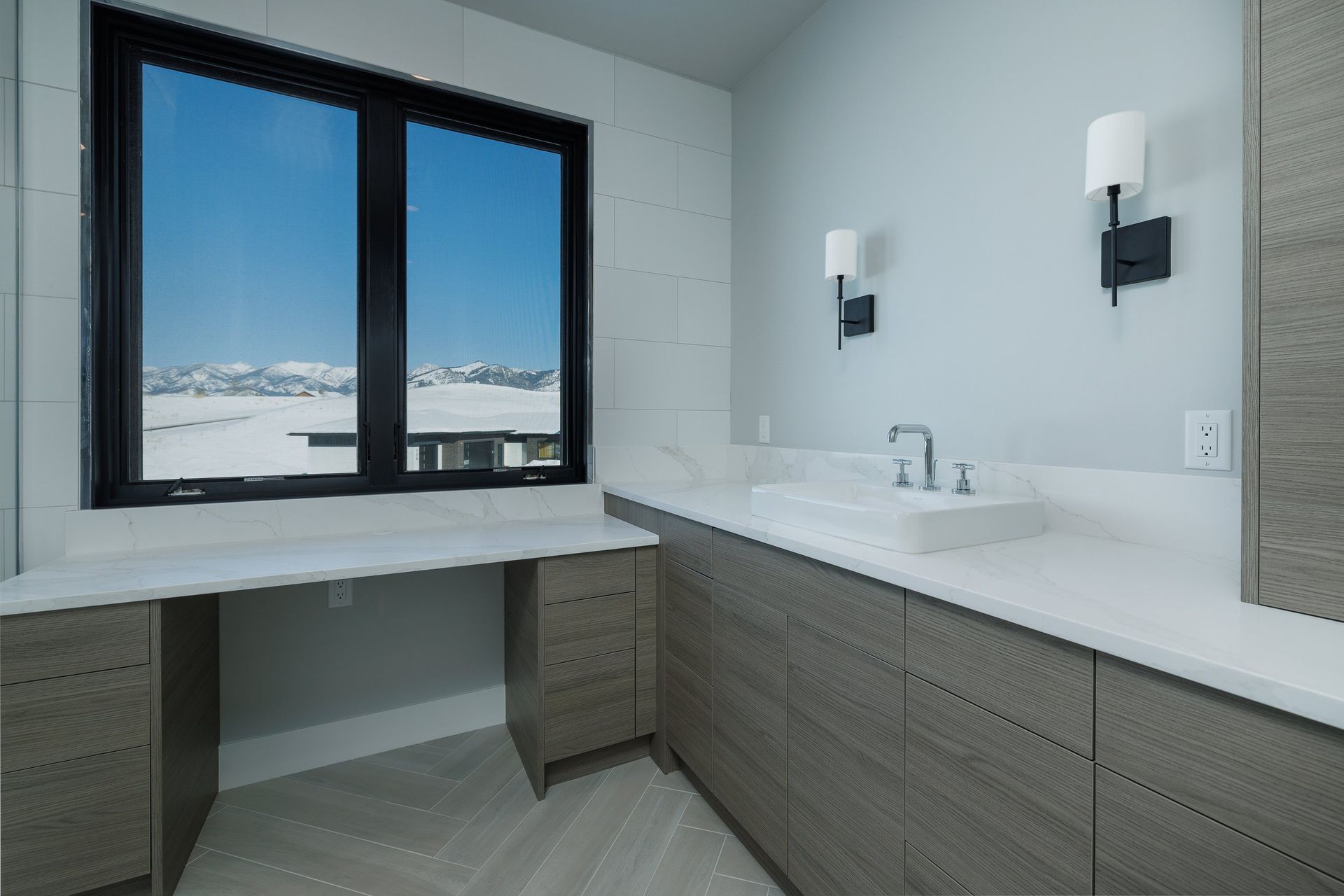 A modern bathroom featuring a white vanity with gray wood cabinets, a sink, wall sconces, and a window with mountain views.