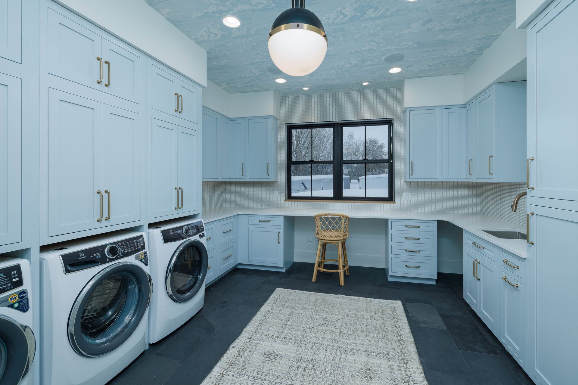 A modern, light blue laundry room with white countertops, dark gray flooring, stacked washers, and a center desk area.