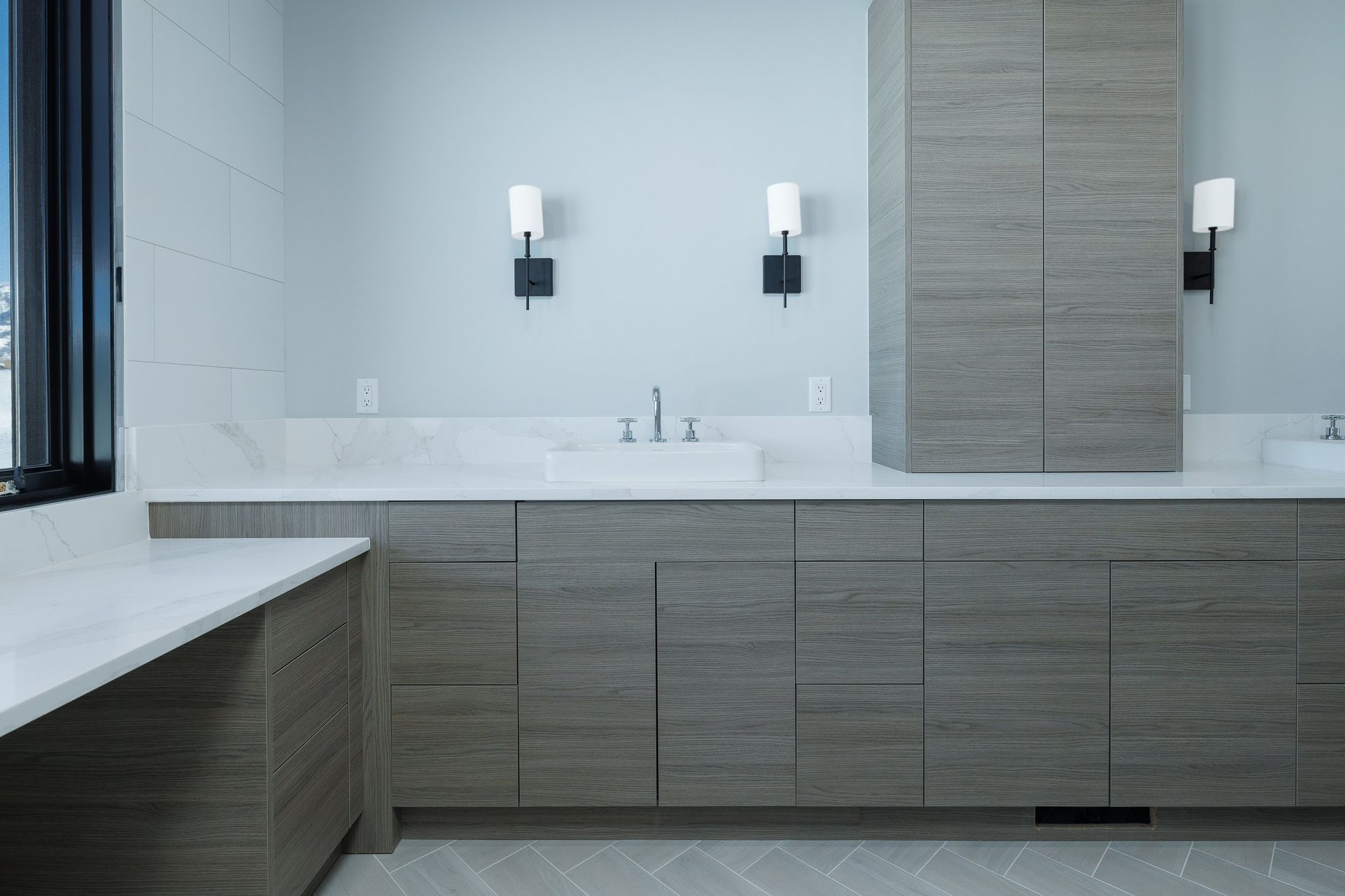 A modern bathroom vanity with gray wood cabinets, white marble countertops, and wall sconces on a light blue wall.