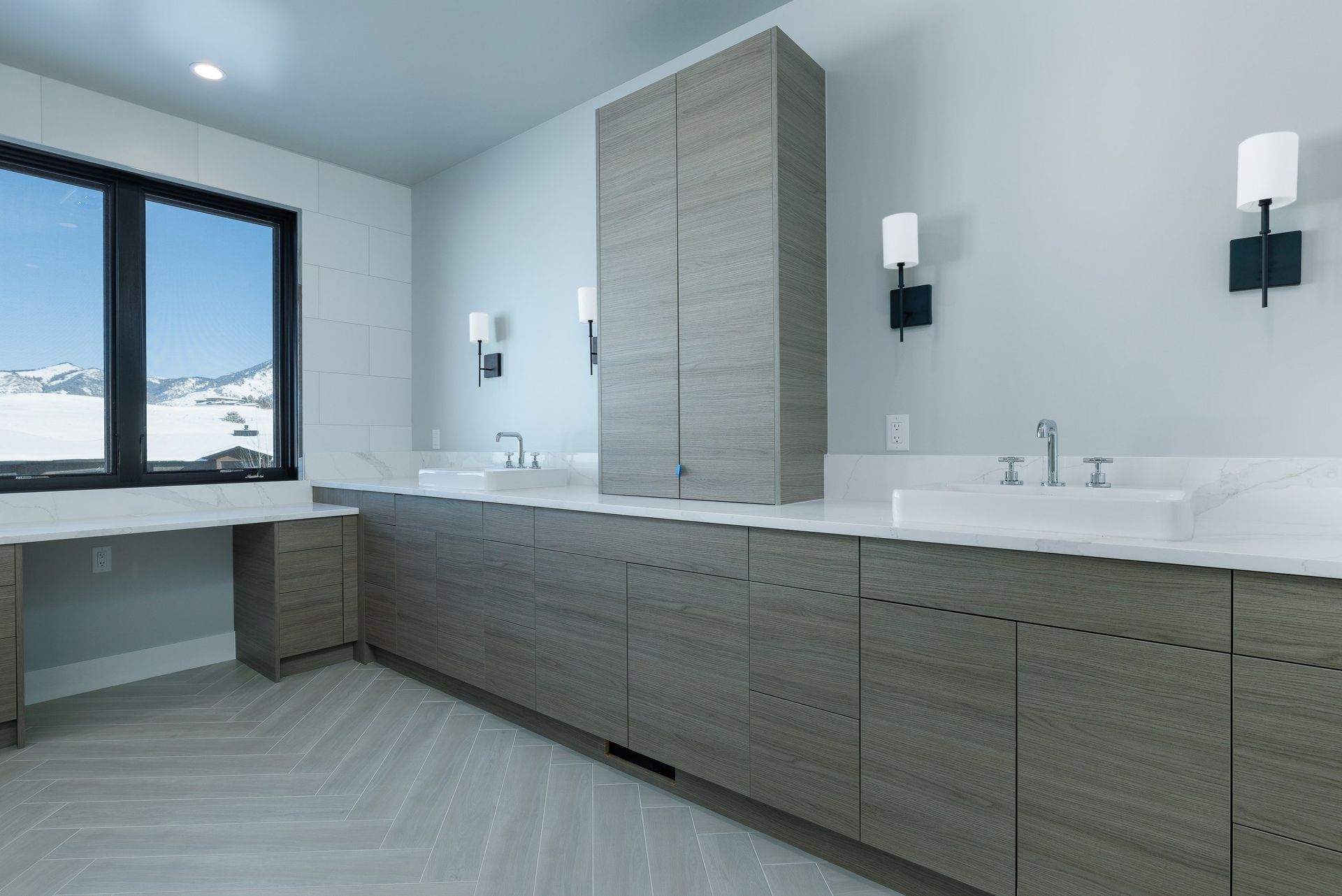Modern bathroom with a long gray wood-grain vanity, two sinks, a tall central cabinet, and a desk area by a large window.