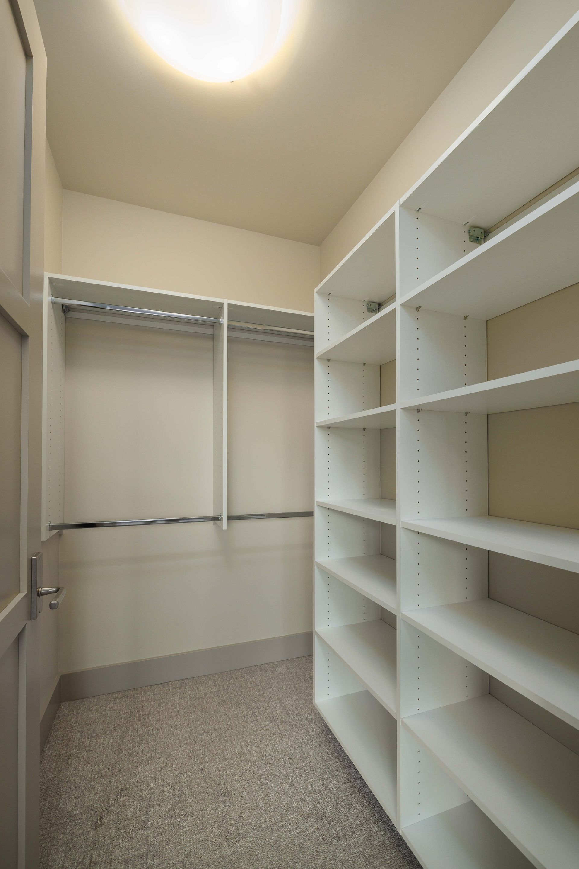 A walk-in closet featuring white wooden shelving units, hanging rods, and a light gray carpeted floor under a ceiling light.