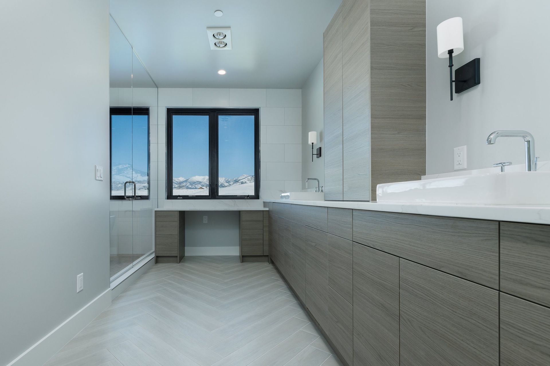 A modern bathroom with grey wood cabinets, a white countertop with two sinks, a herringbone floor, and a large window.