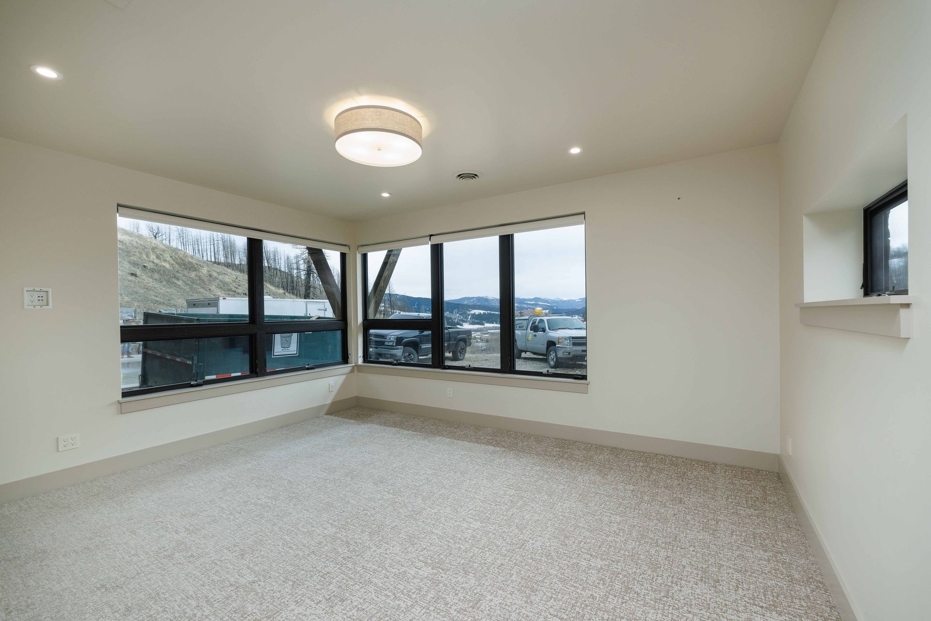 An empty room with light-colored carpet, off-white walls, a central ceiling light, and large windows with black frames.