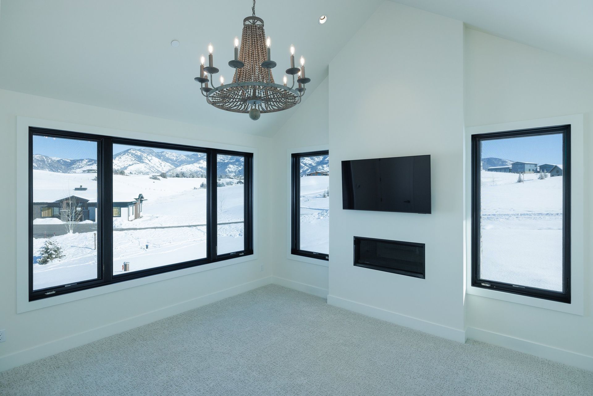 A modern, white room with a chandelier, carpeted floor, fireplace, wall-mounted TV, and large windows with snow views.