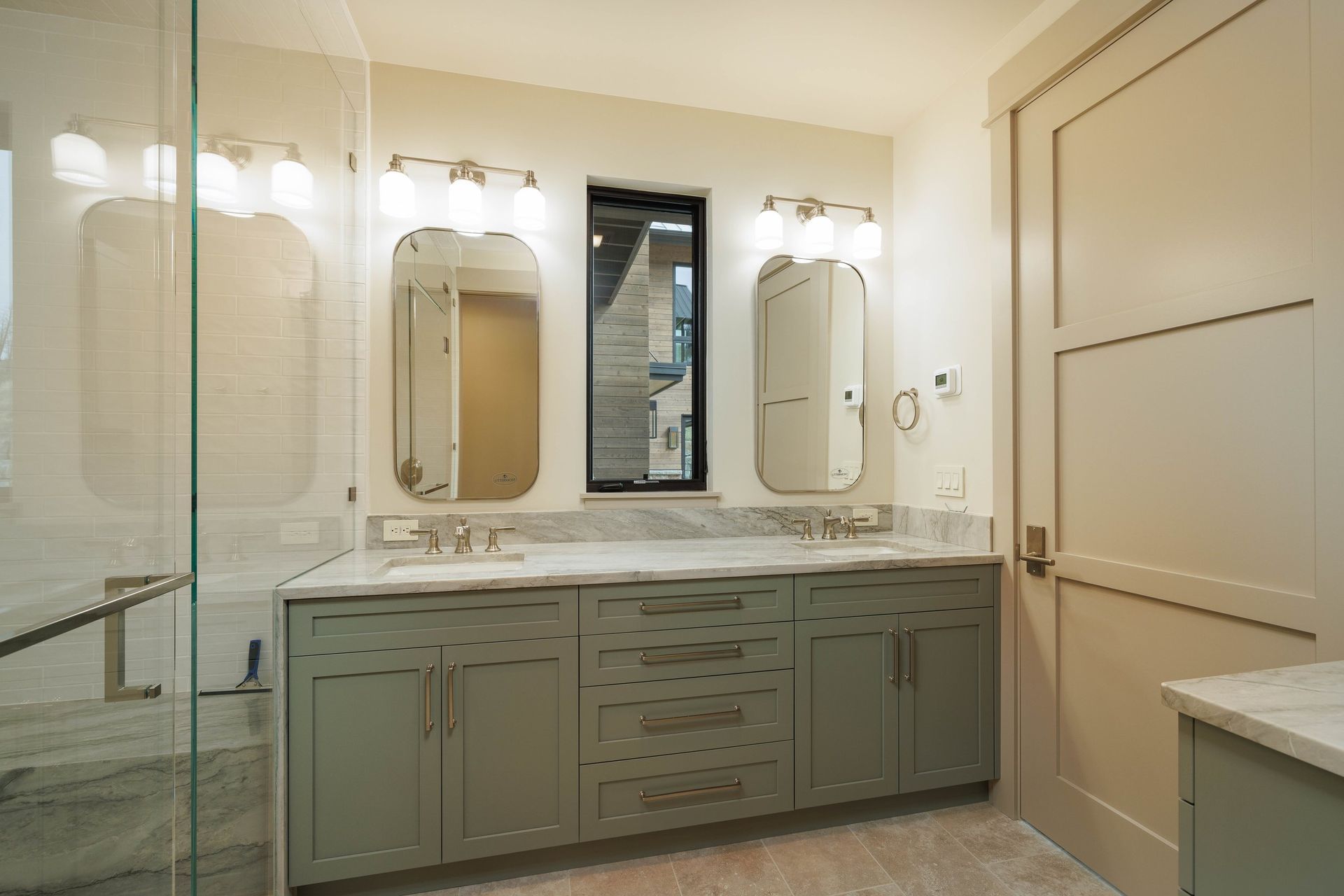 Modern double vanity bathroom with sage green cabinets, marble countertops, two mirrors, and a glass-enclosed shower.