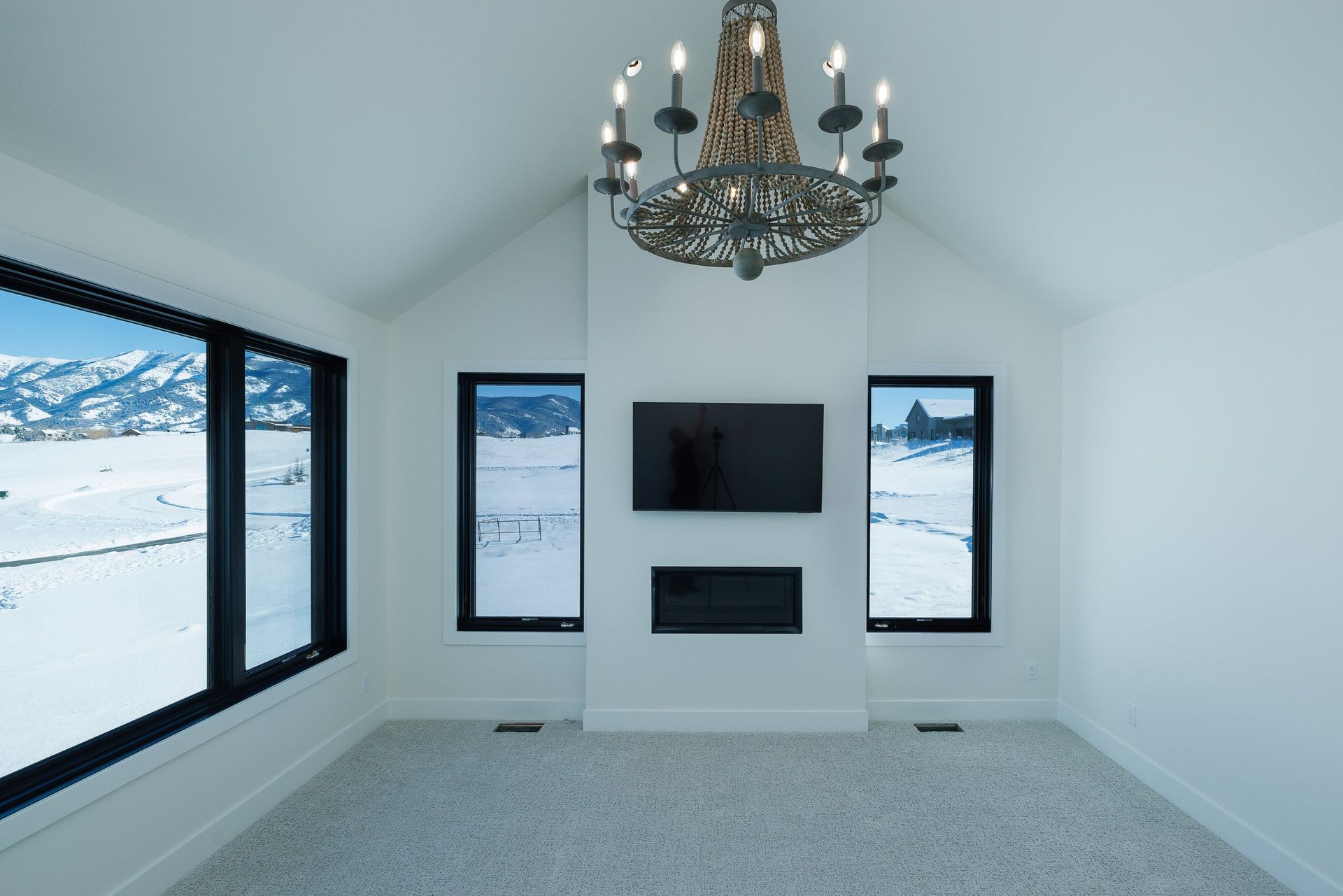 Modern white room with a fireplace, wall-mounted TV, chandelier, and windows showcasing a snowy mountain landscape.