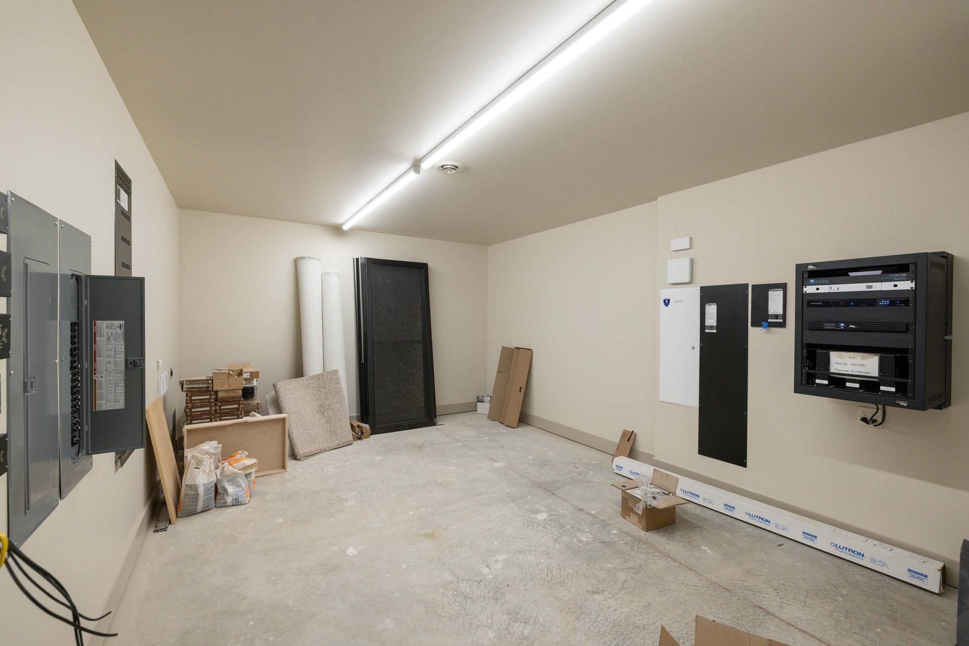 An unfinished, beige-walled storage room with an open electrical panel, cardboard boxes, and a wall-mounted equipment rack.