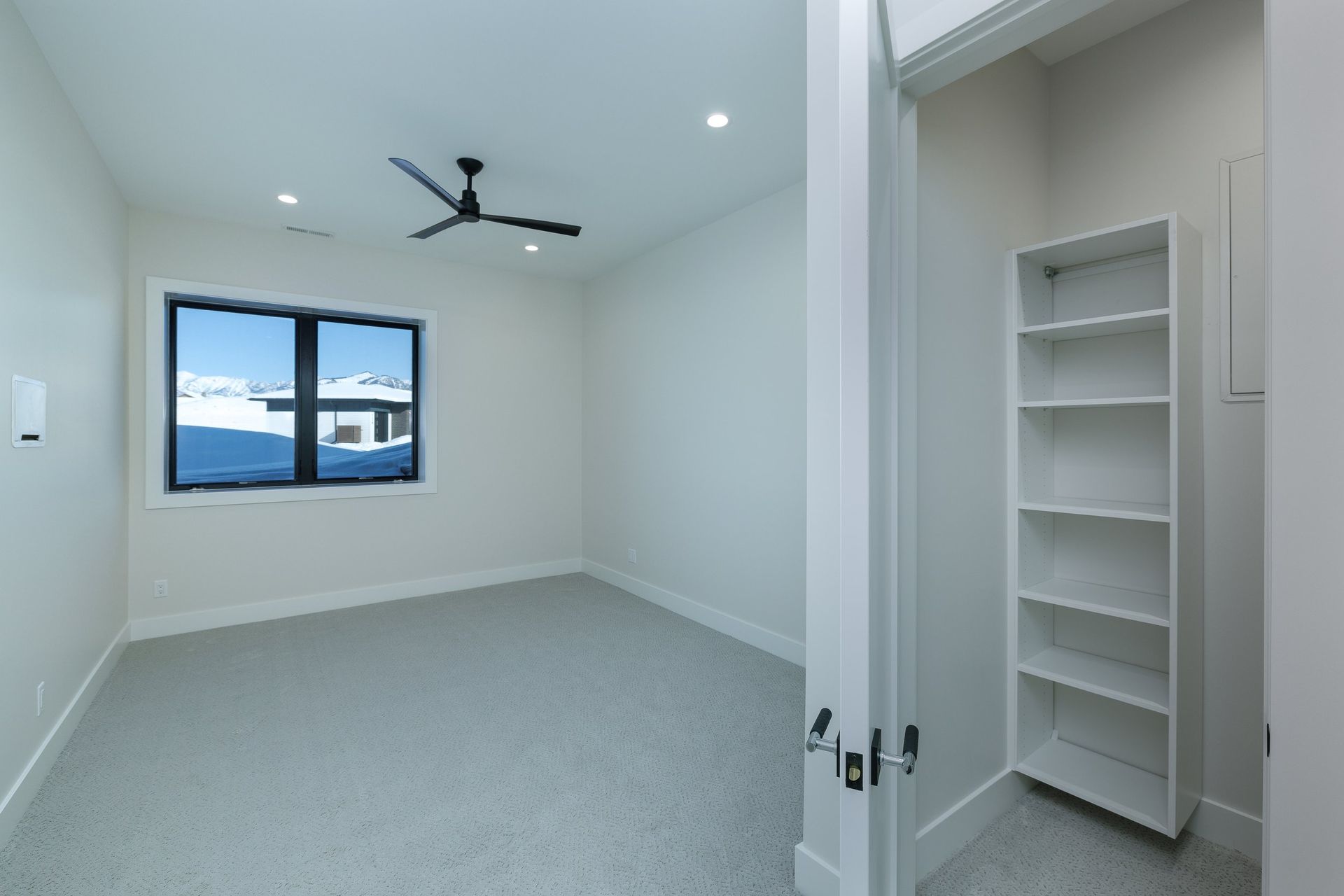A bright, empty bedroom with white walls, gray carpet, a ceiling fan, a window, and a built-in white shelving unit.